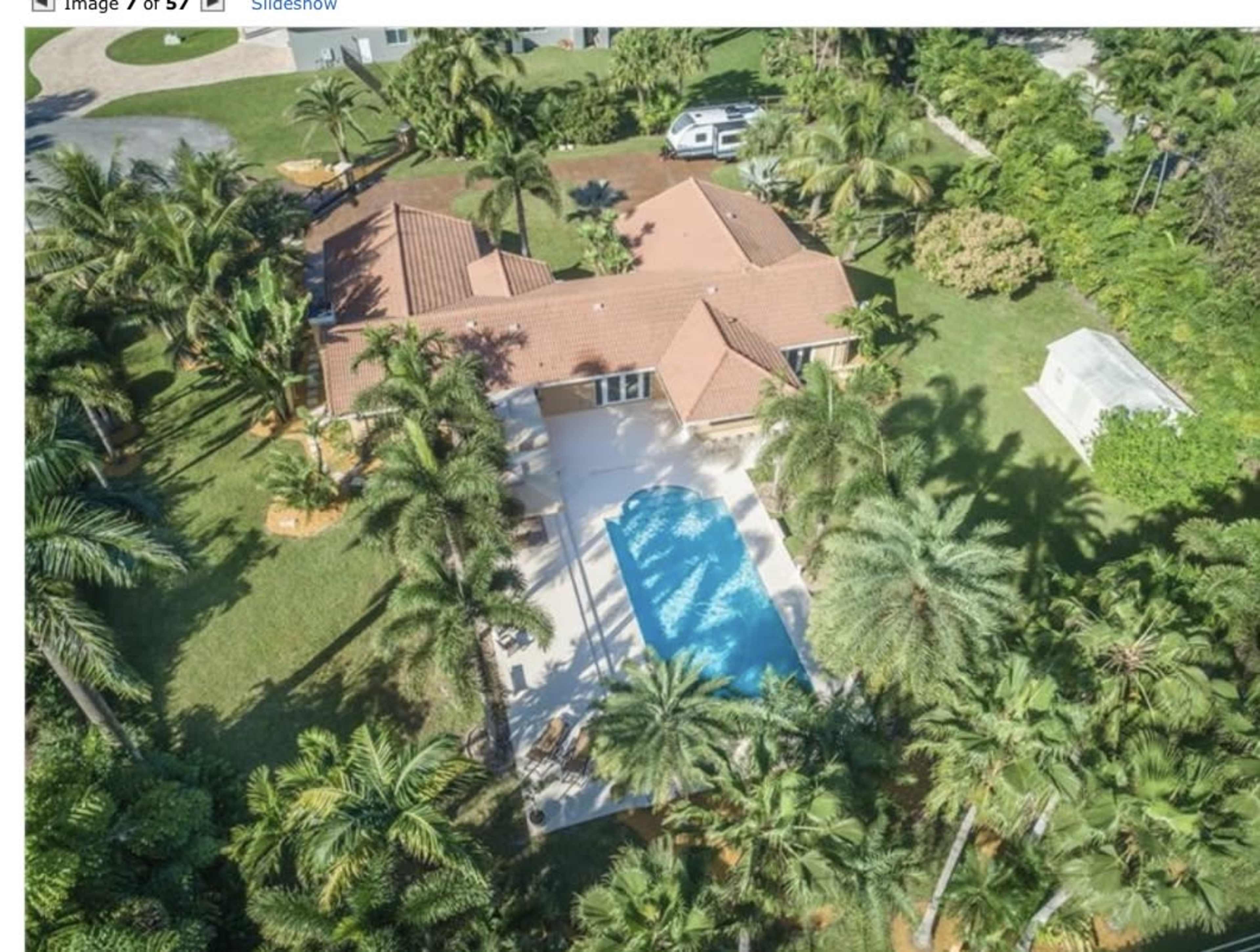 An aerial view shows a house with a pool surrounded by palm trees and lawn in a tropical setting.