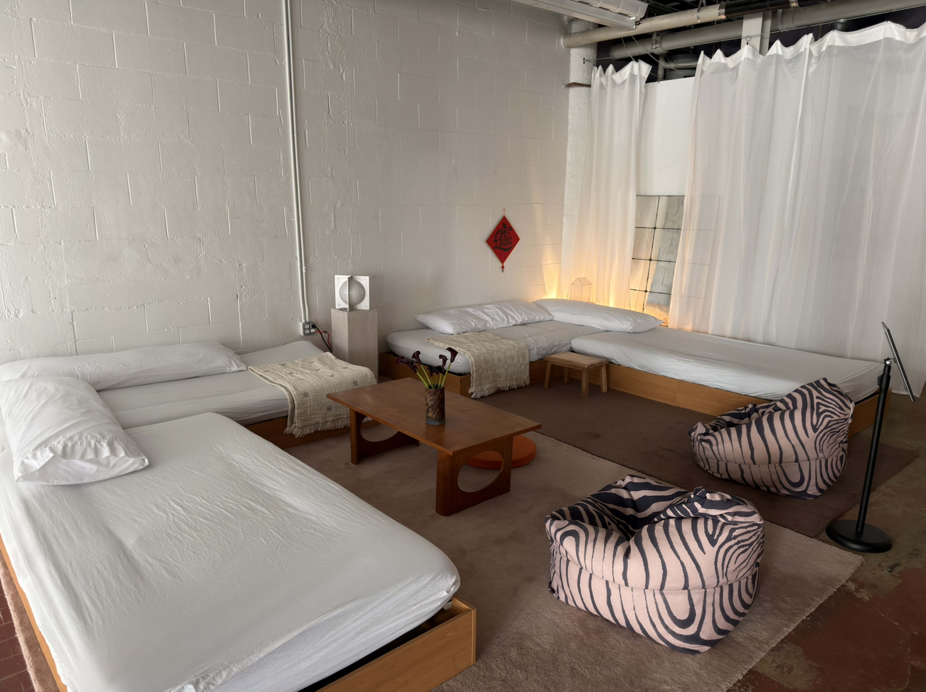 The image depicts a minimalist room featuring several unmade beds, a wooden table in the center, and two beanbag chairs, all arranged around a softly lit area with sheer curtains.
