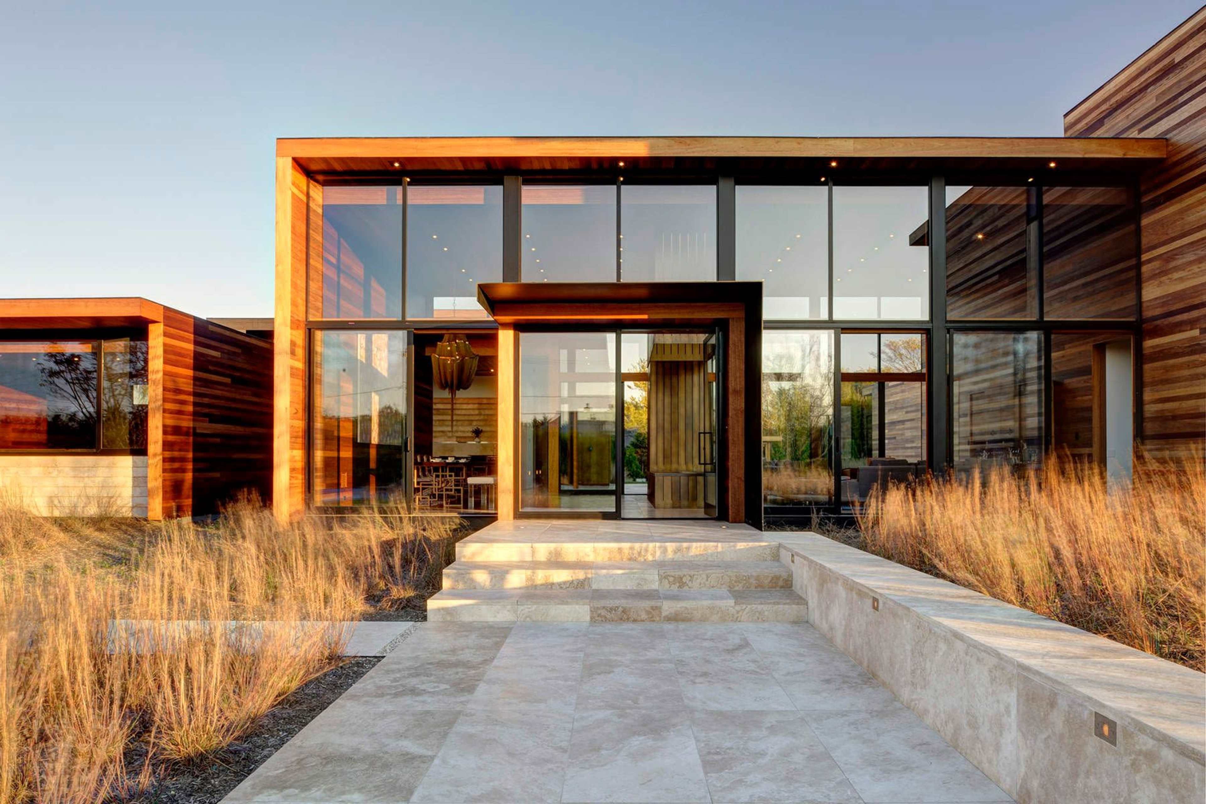 A modern house with large glass windows and wooden accents, set against a backdrop of wild grass and a clear sky.