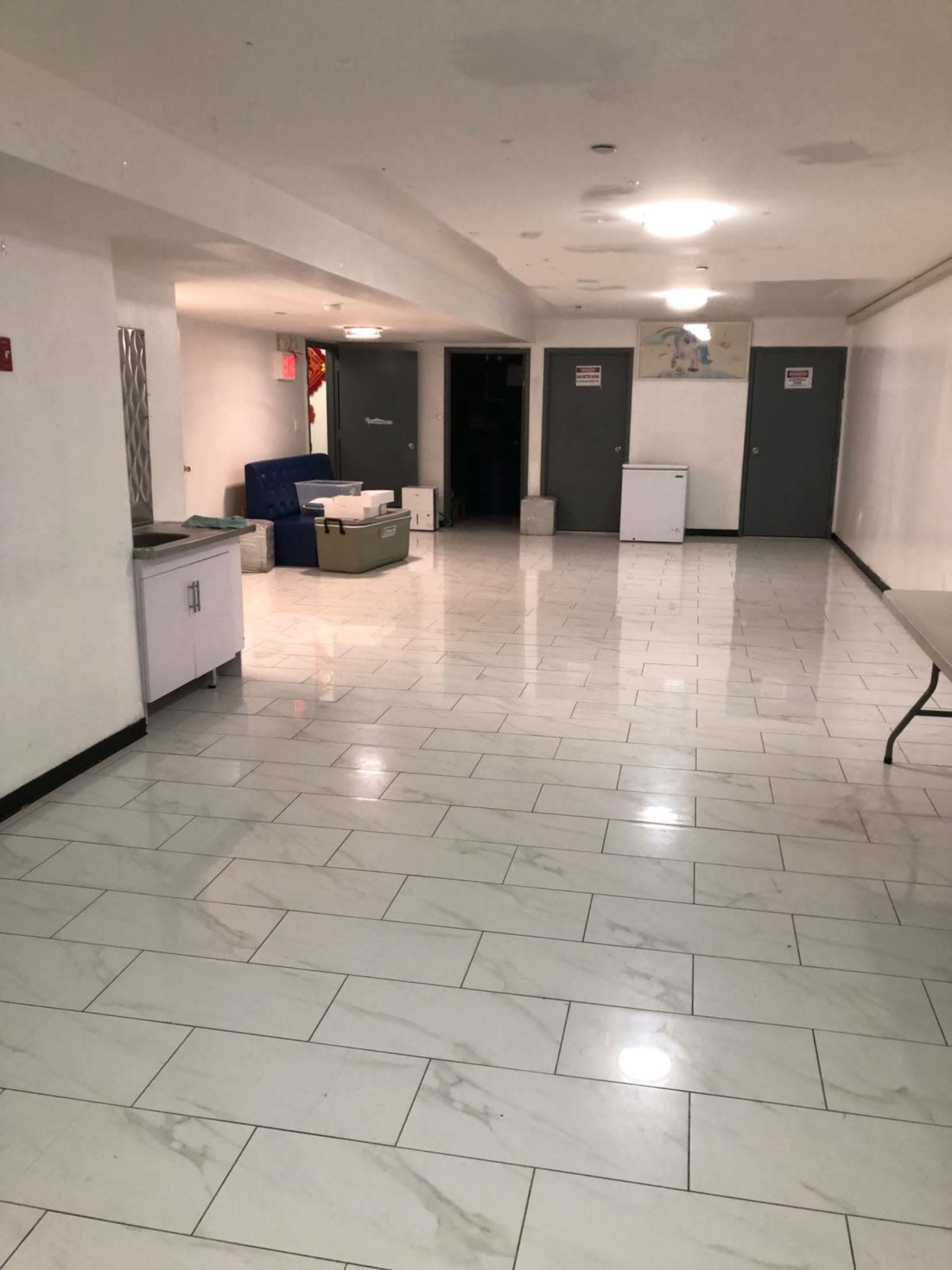 The image shows a spacious, empty indoor area with white tiled floors, several gray doors, and minimal furnishings including a table and some seating.