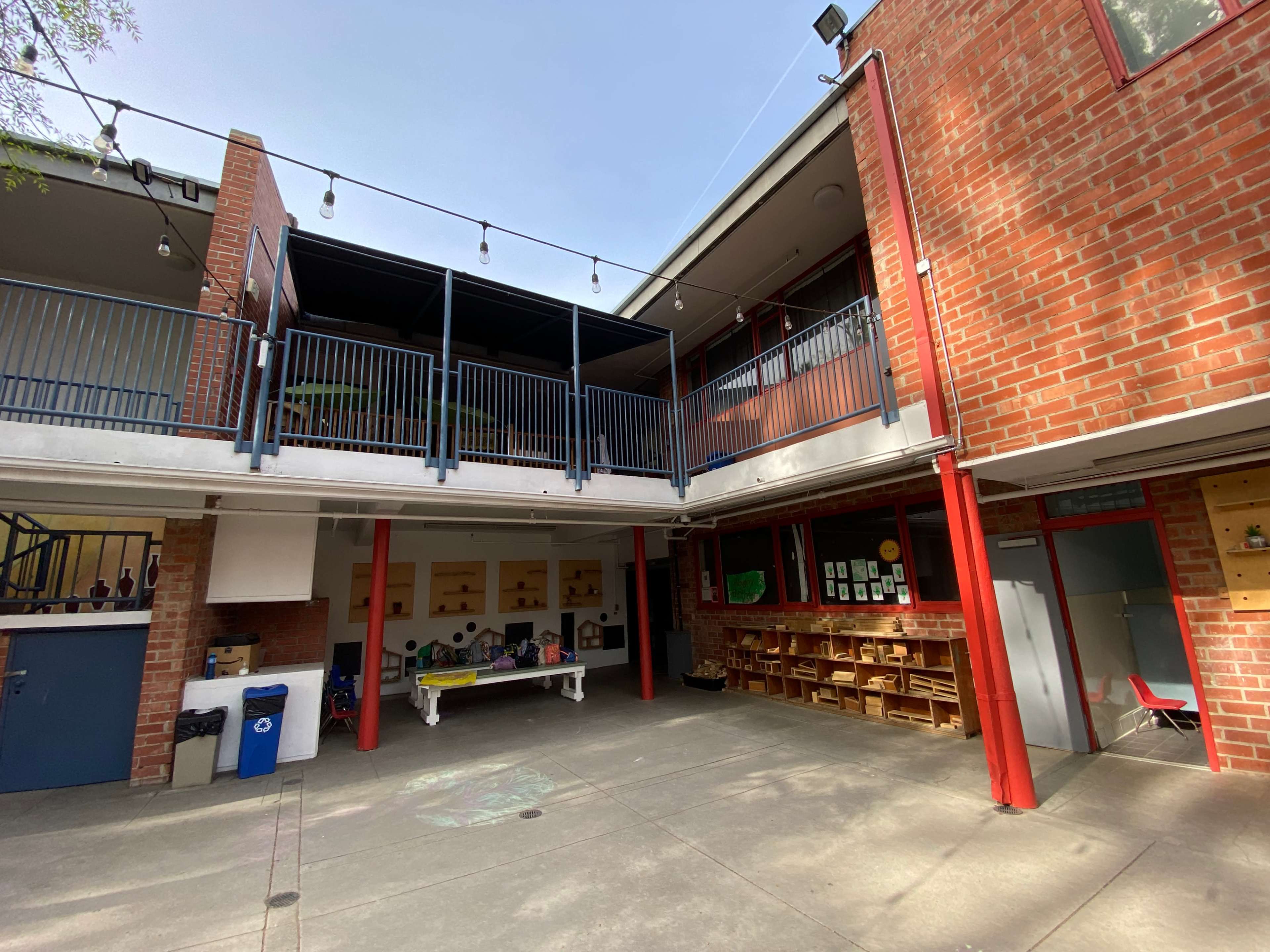 A courtyard area of a school building with a brick exterior, featuring a two-story structure and tables arranged beneath an overhang.