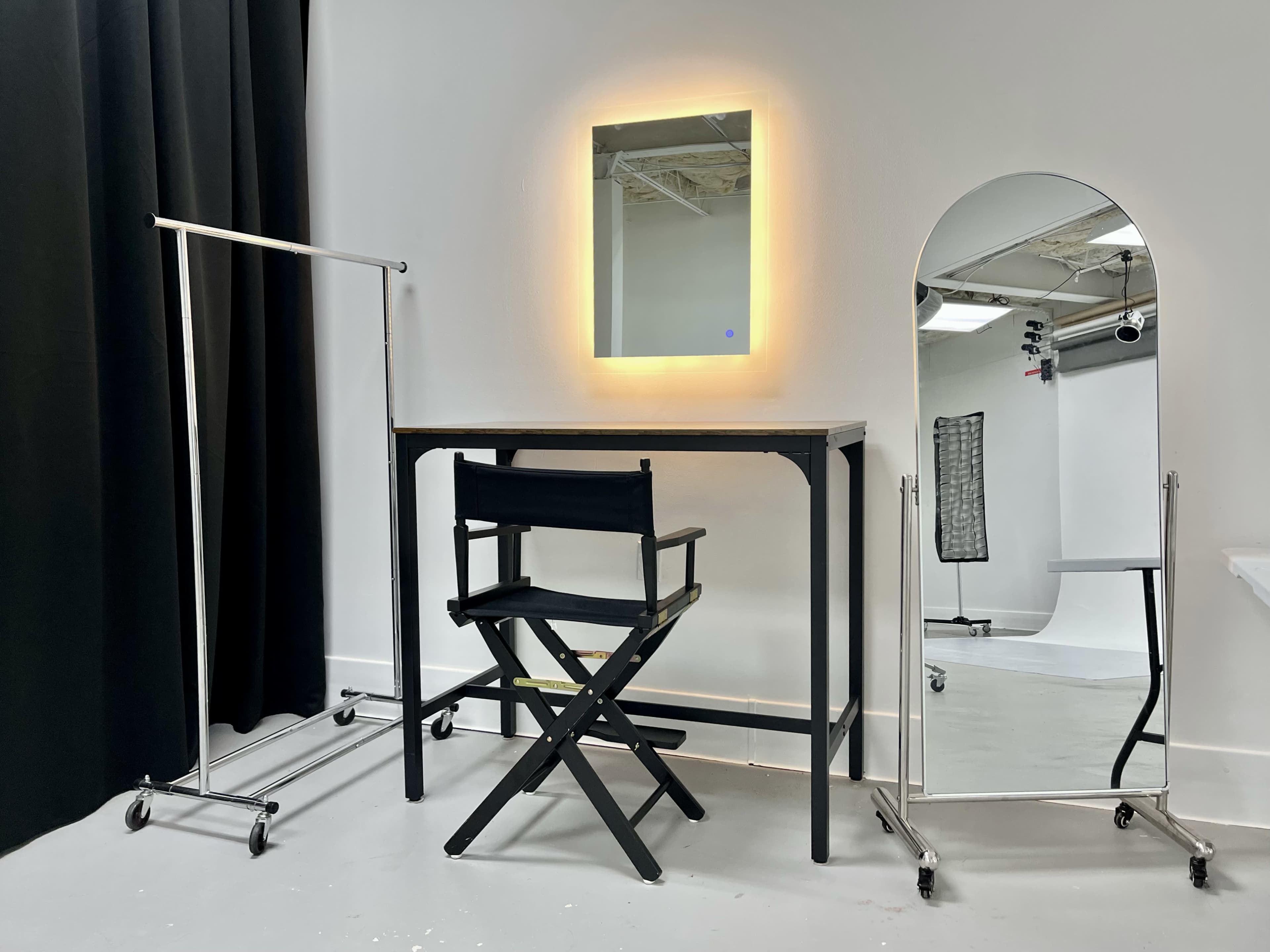 The image shows a minimalist dressing area featuring a black director's chair, a wooden table with a backlit mirror, and a tall, arched mirror beside a clothing rack on wheels.