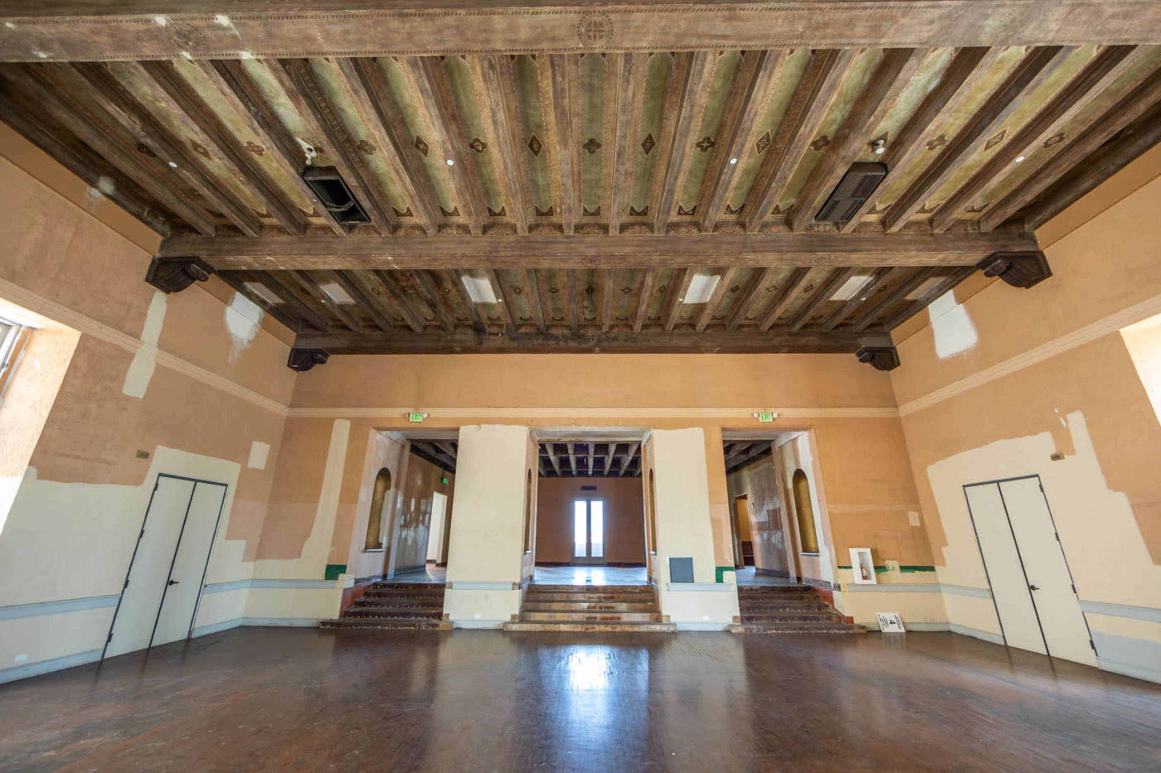The image shows a large, empty room with a wooden beam ceiling and multiple doorways leading to a bright area at the far end.