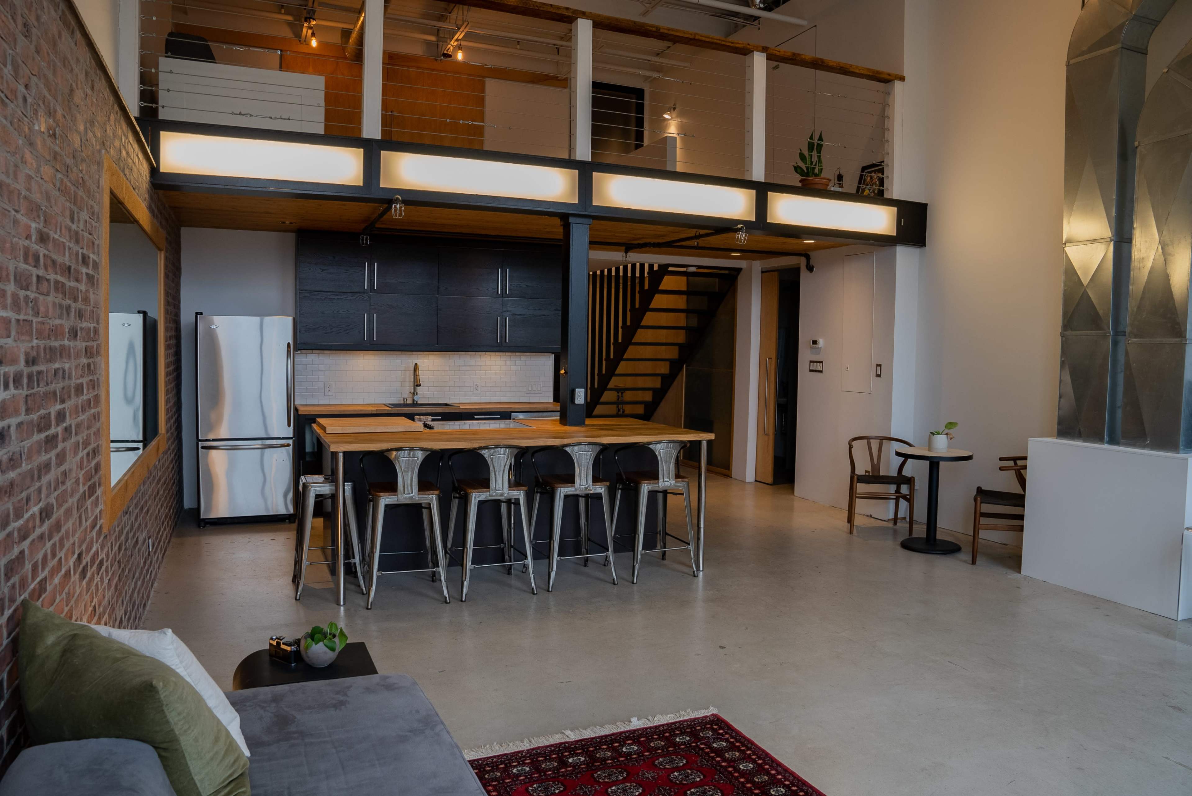 A modern open-concept loft features a kitchen island with barstools, a dining table, and a staircase leading to a mezzanine level.