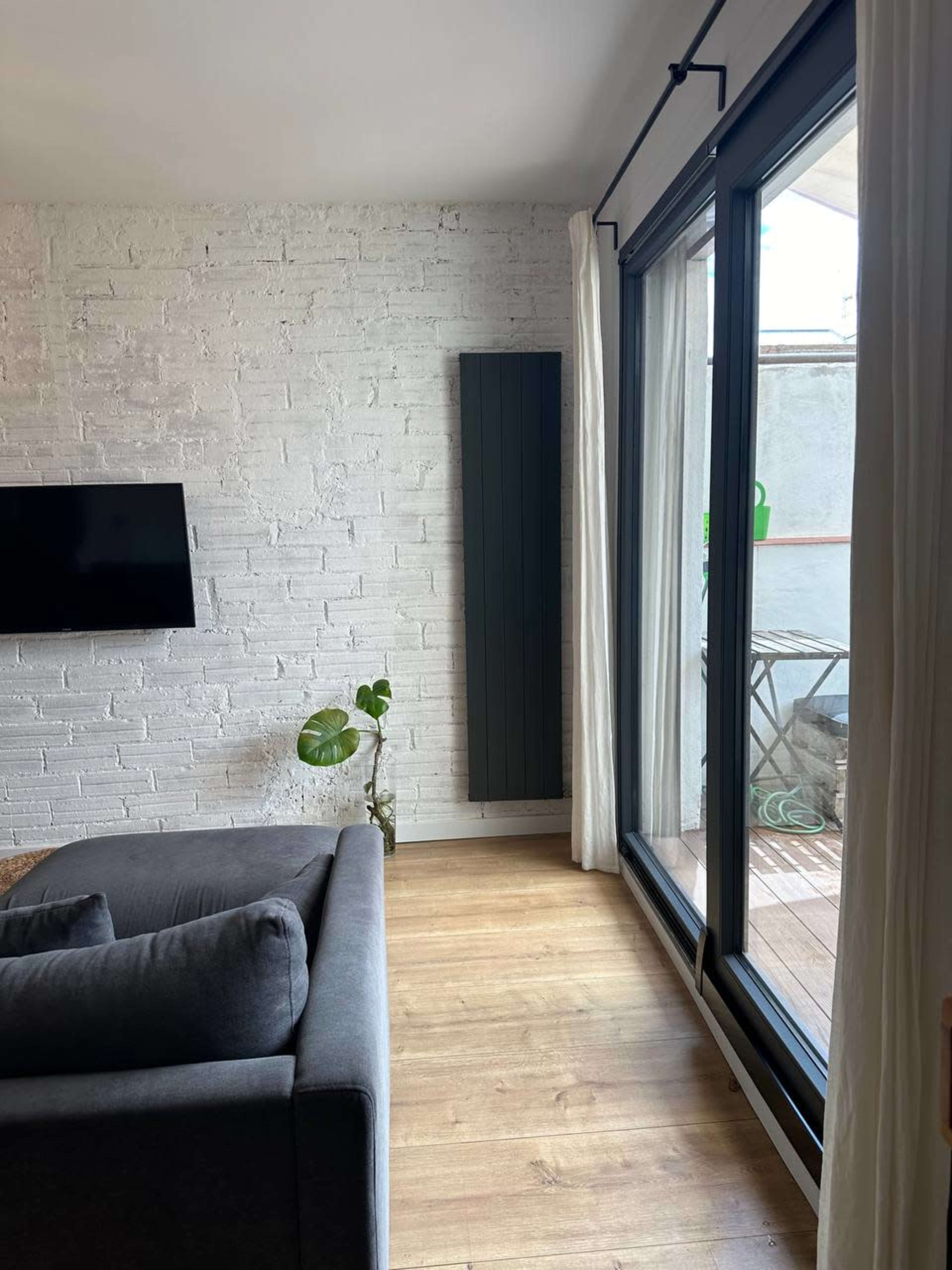 A modern living room features a gray sofa, a white brick wall, a mounted television, and a large window with sliding glass doors.