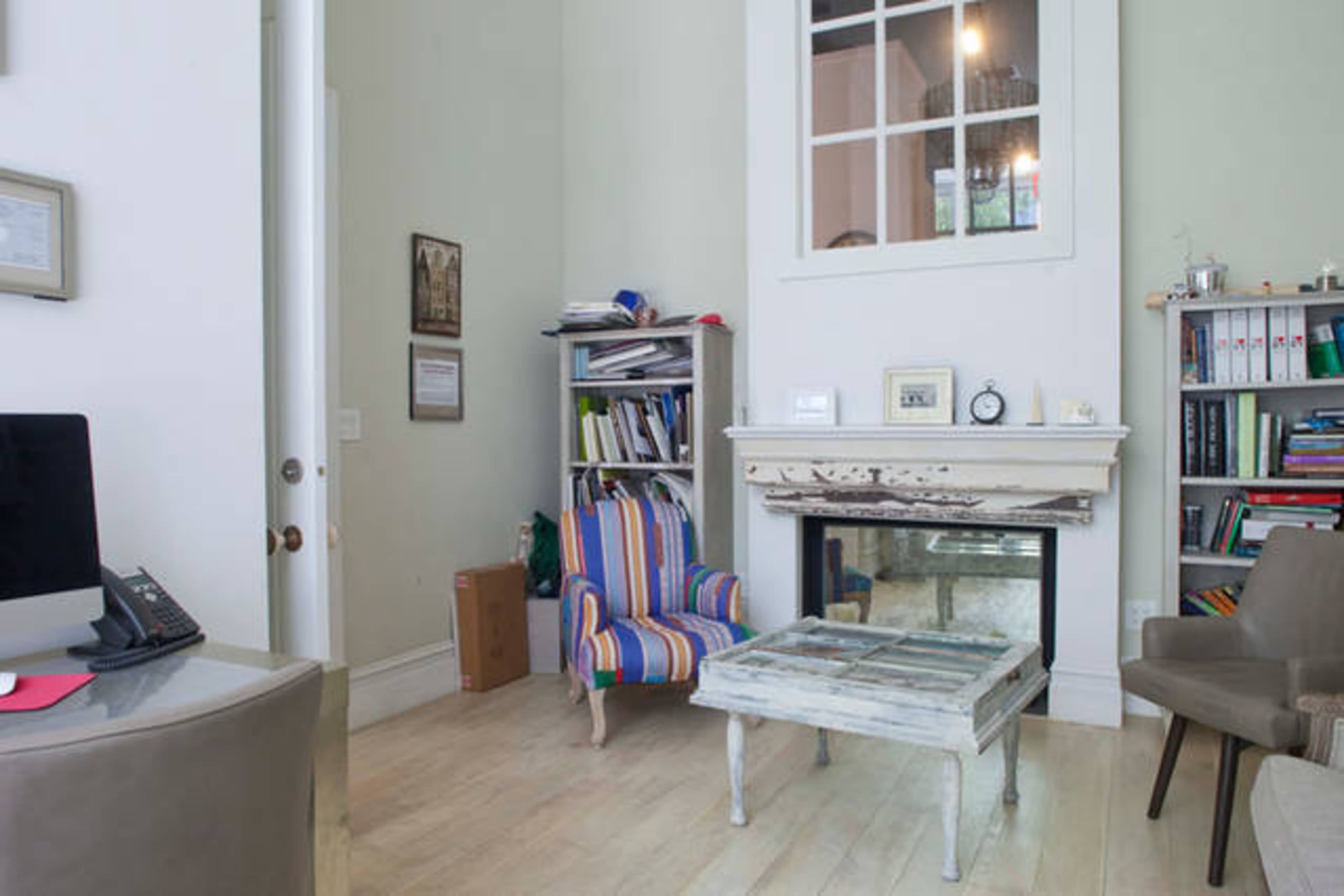 A small room features a colorful striped armchair, a glass-topped coffee table, a decorative fireplace, and a bookshelf filled with books.