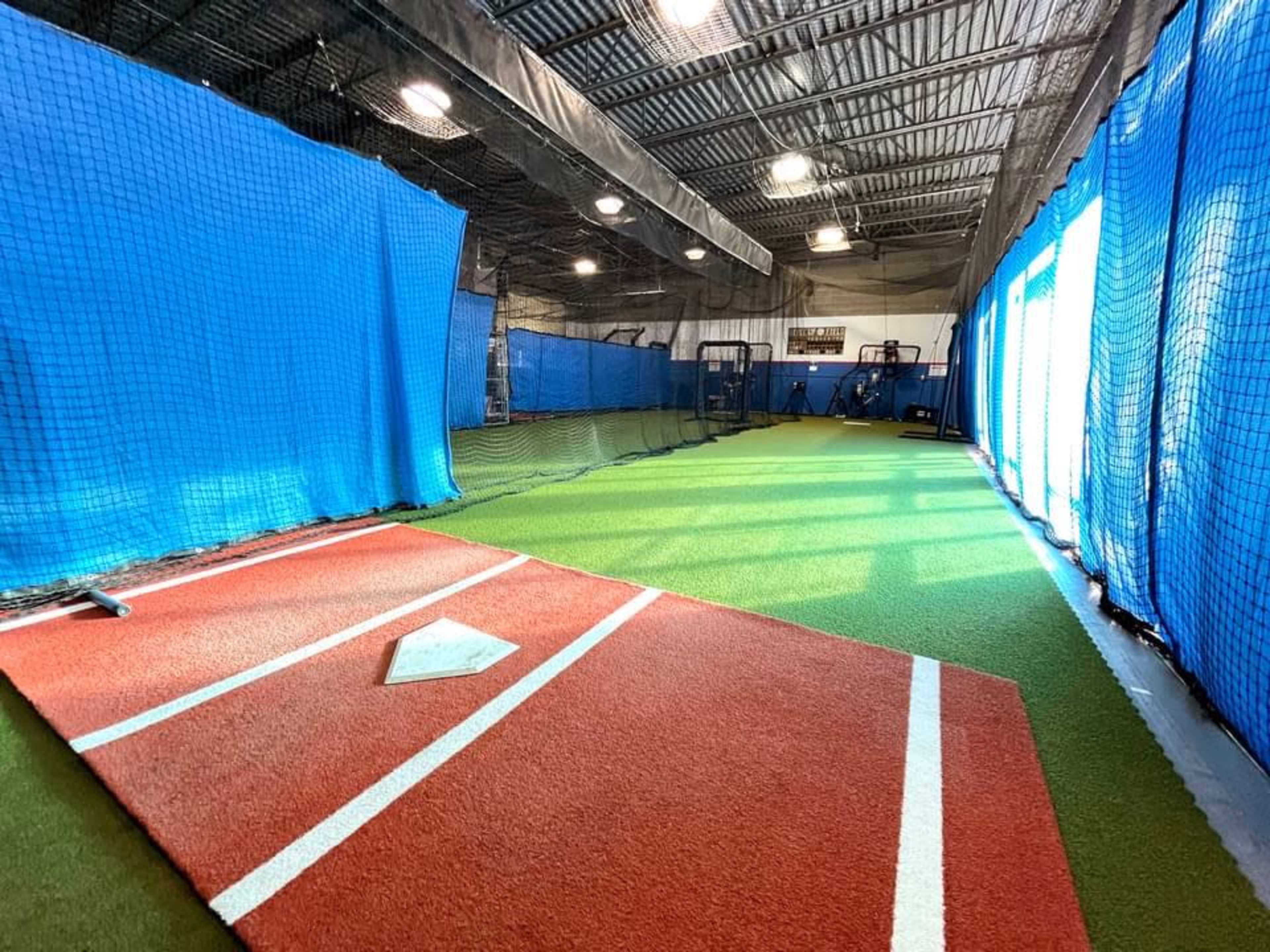 An indoor sports facility featuring a baseball practice area with a red pitching mound and blue netting partitions.