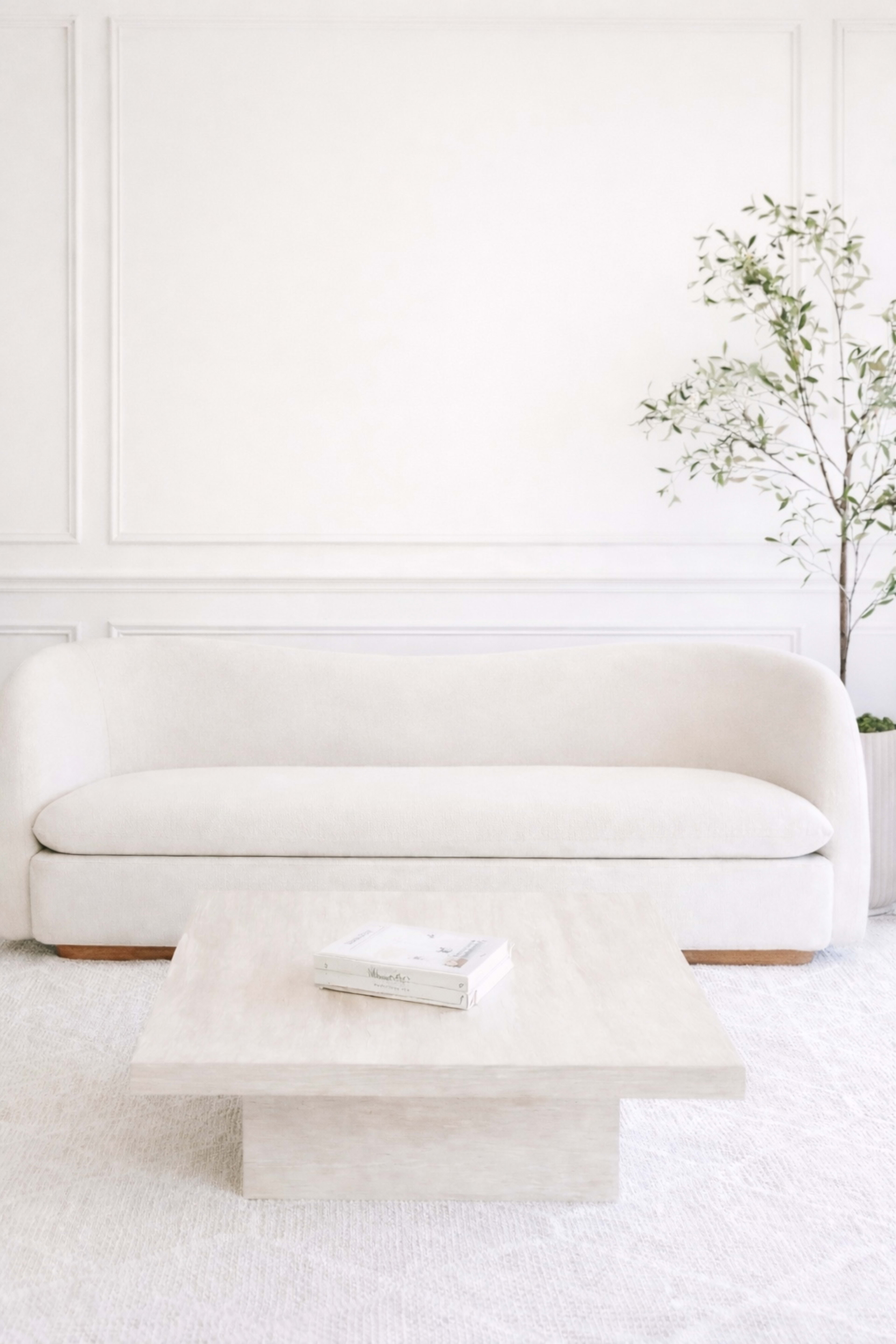 A light-colored sofa is positioned in front of a square coffee table, set against a plain white wall, with an indoor plant in the corner.