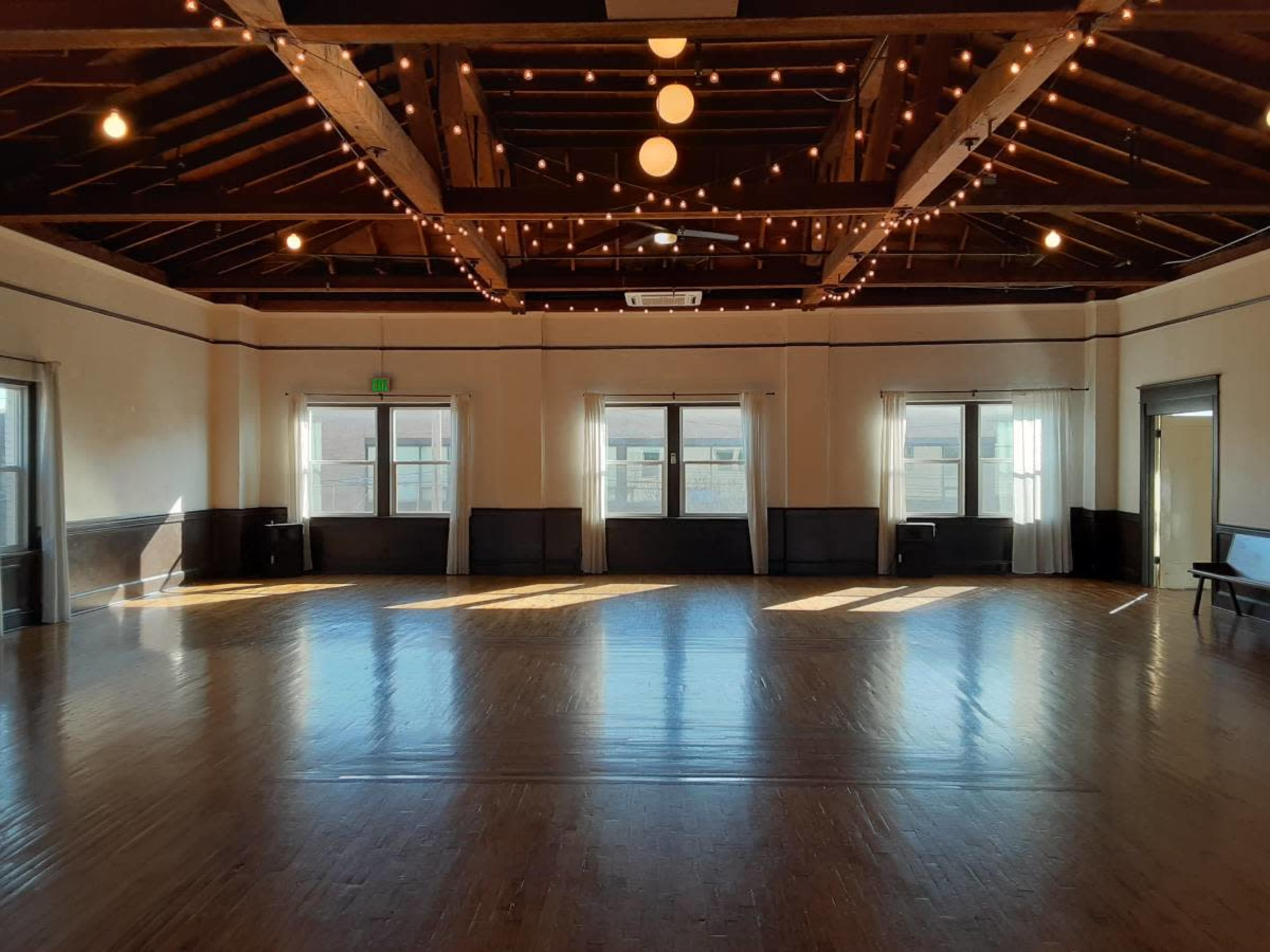 A spacious wooden dance studio with large windows allowing natural light, string lights along the ceiling, and polished hardwood floors.