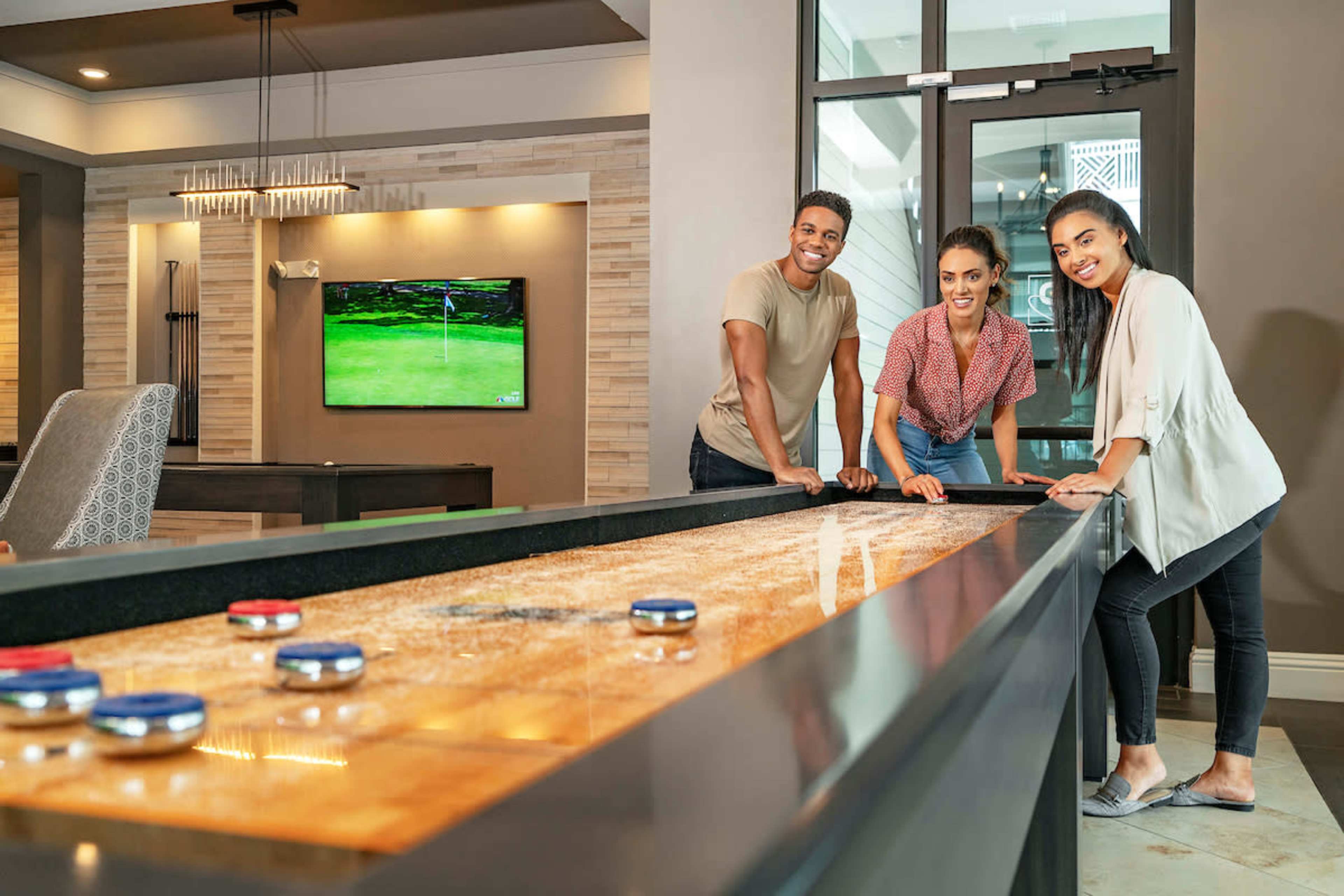 Three people are smiling and leaning on a shuffleboard table in a modern common area with a television displaying a golf game in the background.