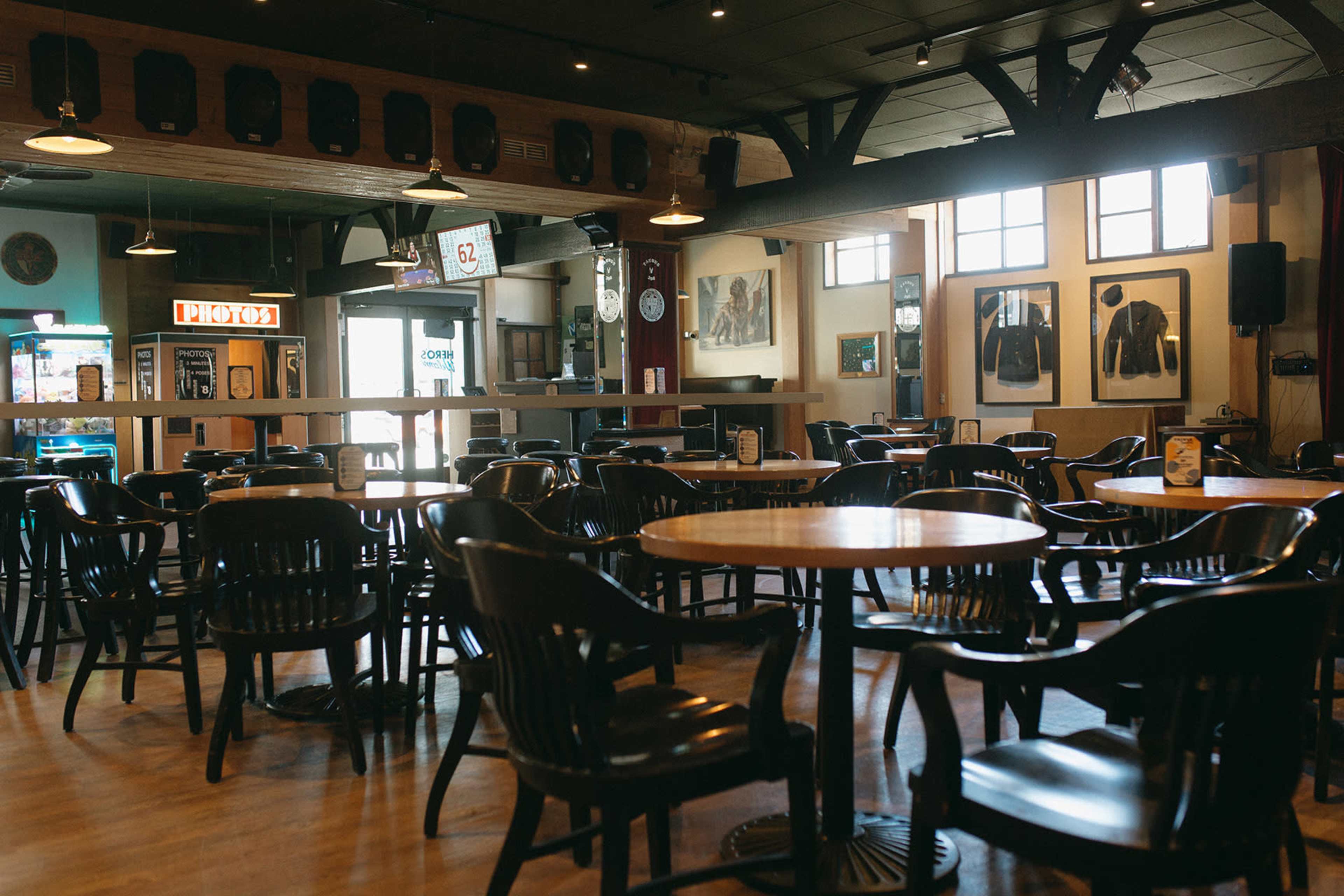 The interior of a restaurant features empty wooden tables and chairs, with large windows and framed artwork on the walls.