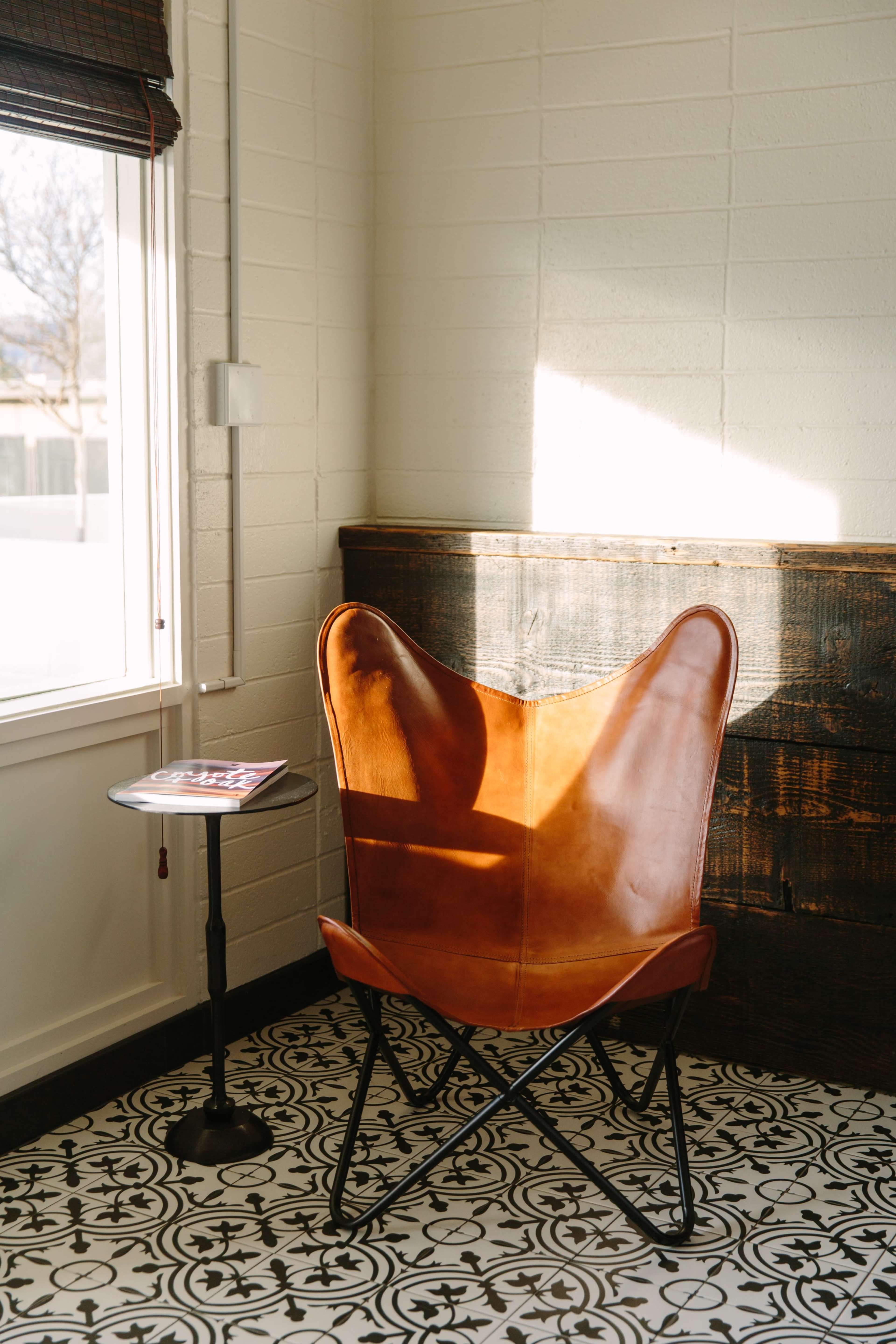 A leather butterfly chair sits beside a small table in a brightly lit corner of a room with patterned tile flooring and a wooden wall.