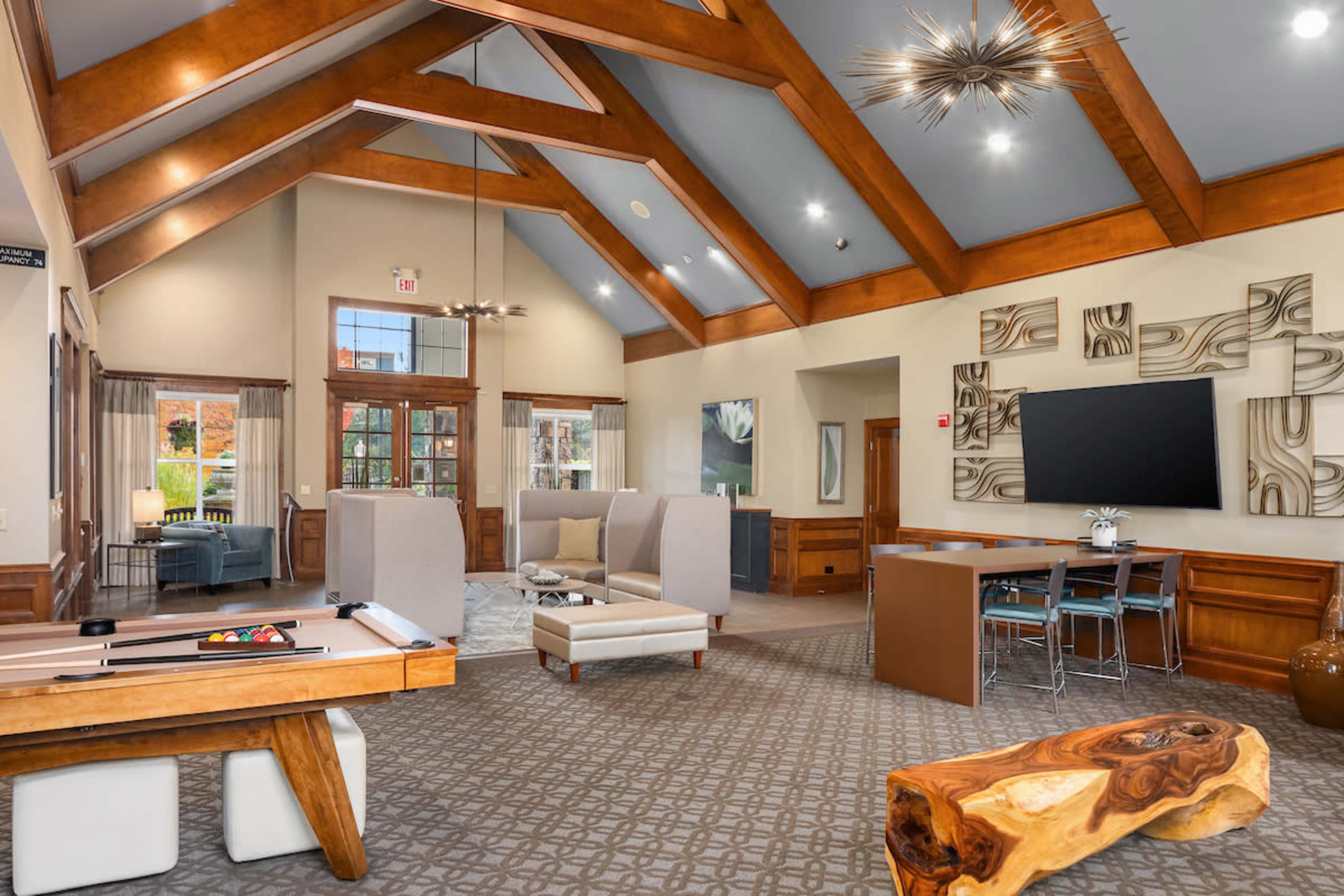 The image features a spacious lobby area with wooden beams, modern seating arrangements, and a pool table, surrounded by large windows and contemporary decor.