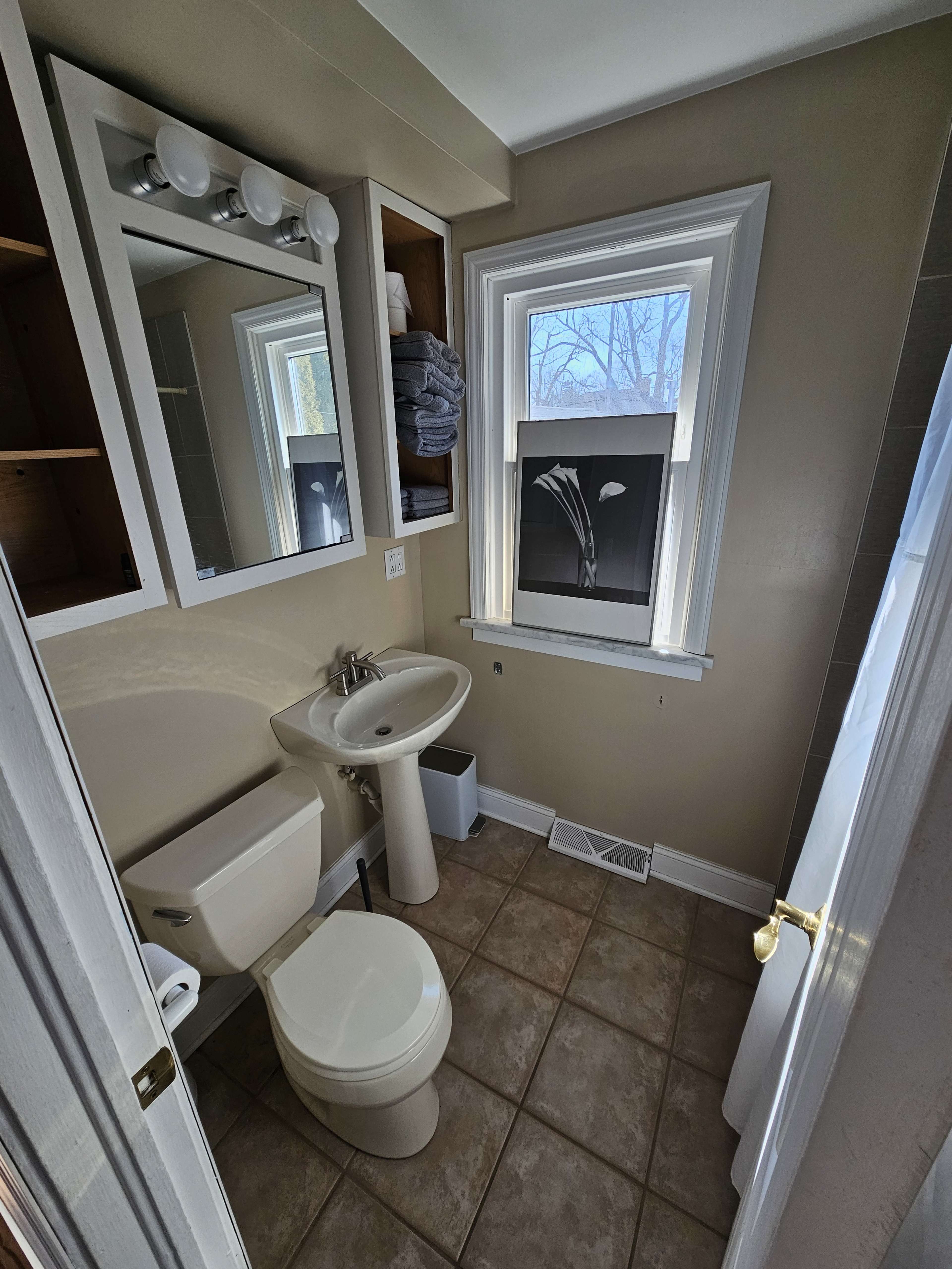A small bathroom featuring a toilet, sink, and a window, with shelves holding towels and a framed picture on the wall.
