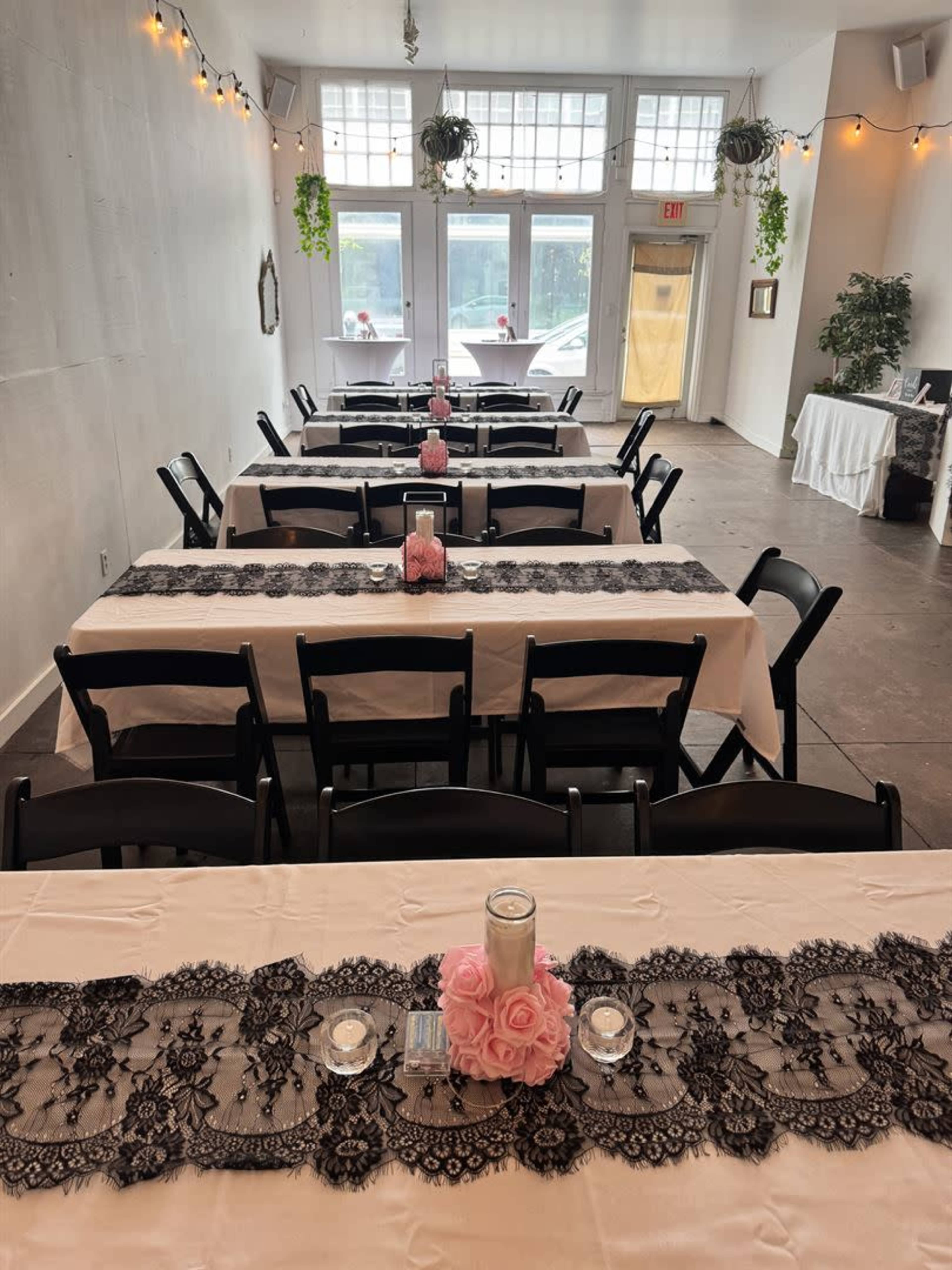 A decorative banquet room features several long tables set with lace runners, candles, and pink roses.