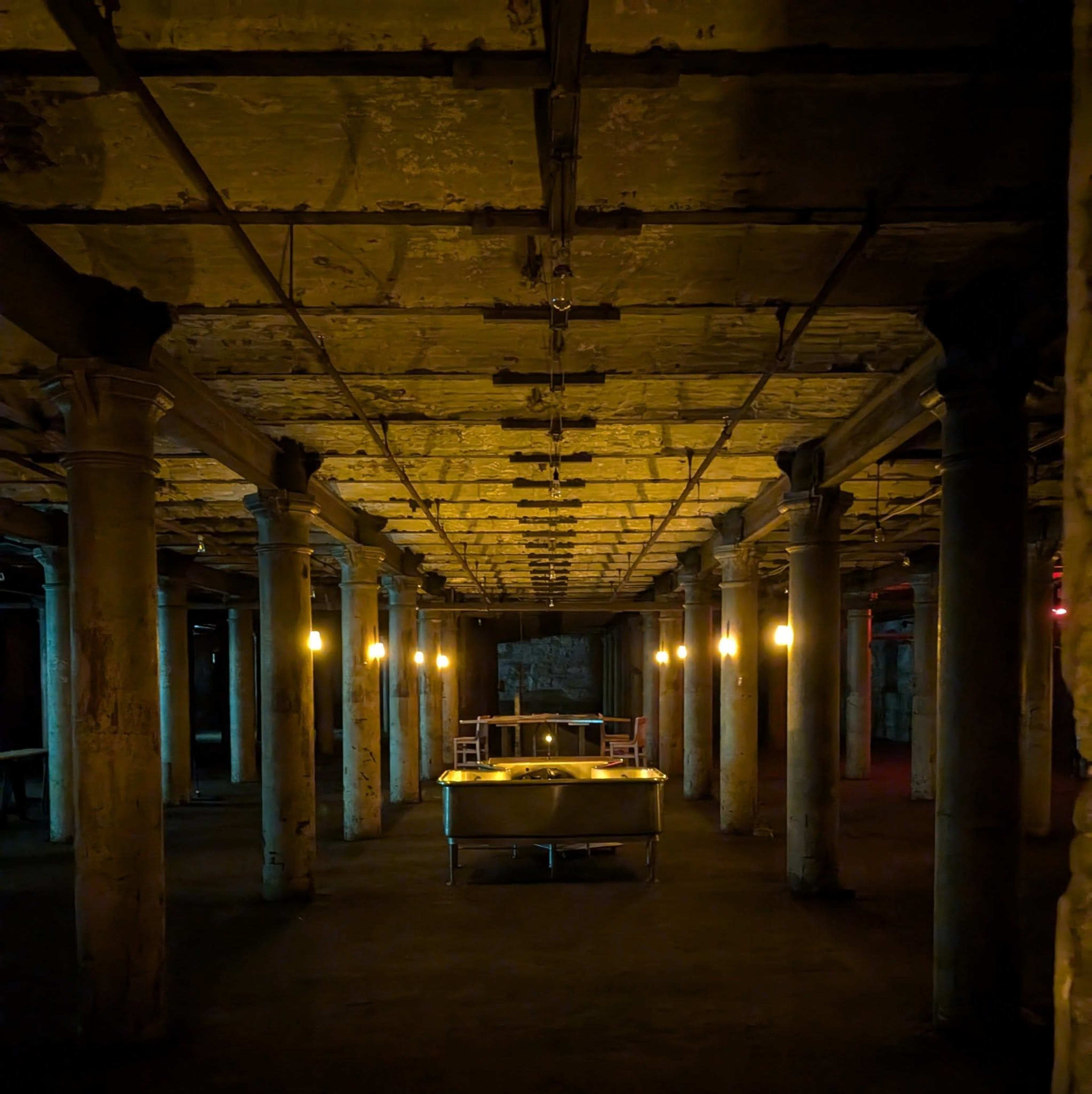 The image shows a dimly lit basement with stone pillars, a long table at the center, and soft overhead lighting creating shadows against the textured walls and ceiling.