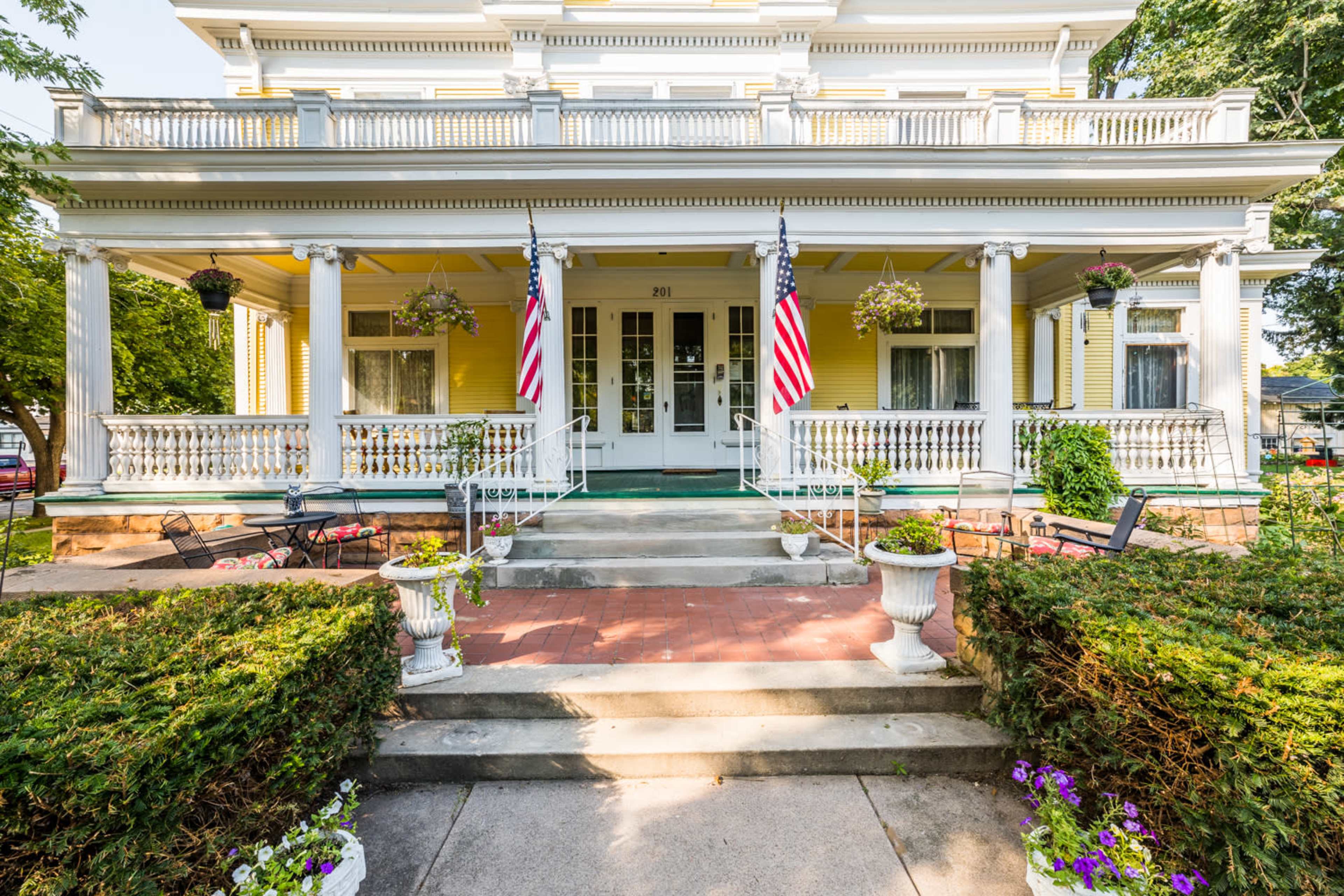 A large yellow Victorian-style house with a welcoming porch, flanked by potted plants and two American flags.