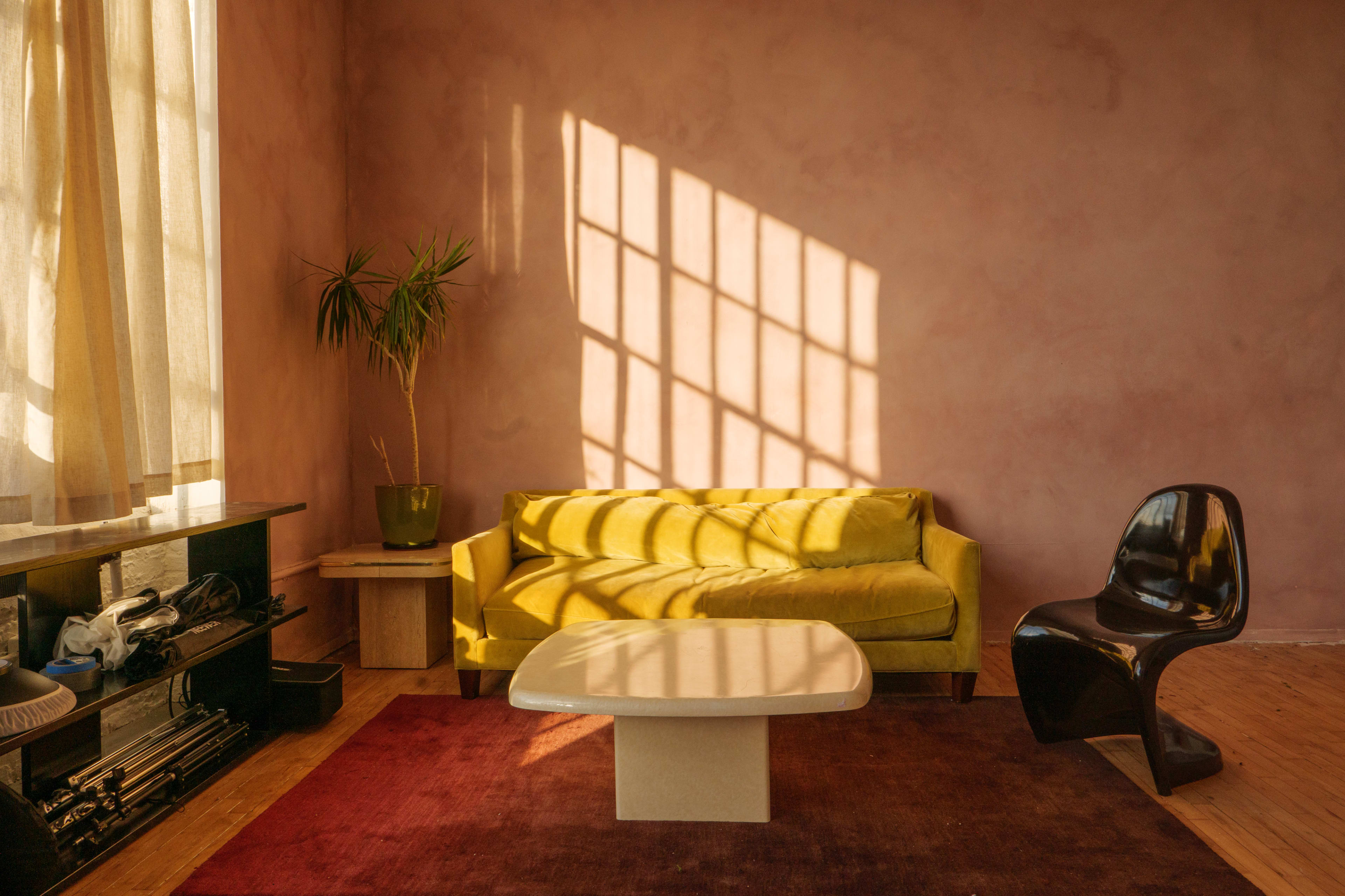 A yellow sofa and a black chair are positioned in a room with pink walls, where sunlight casts long shadows through a grid-patterned window.