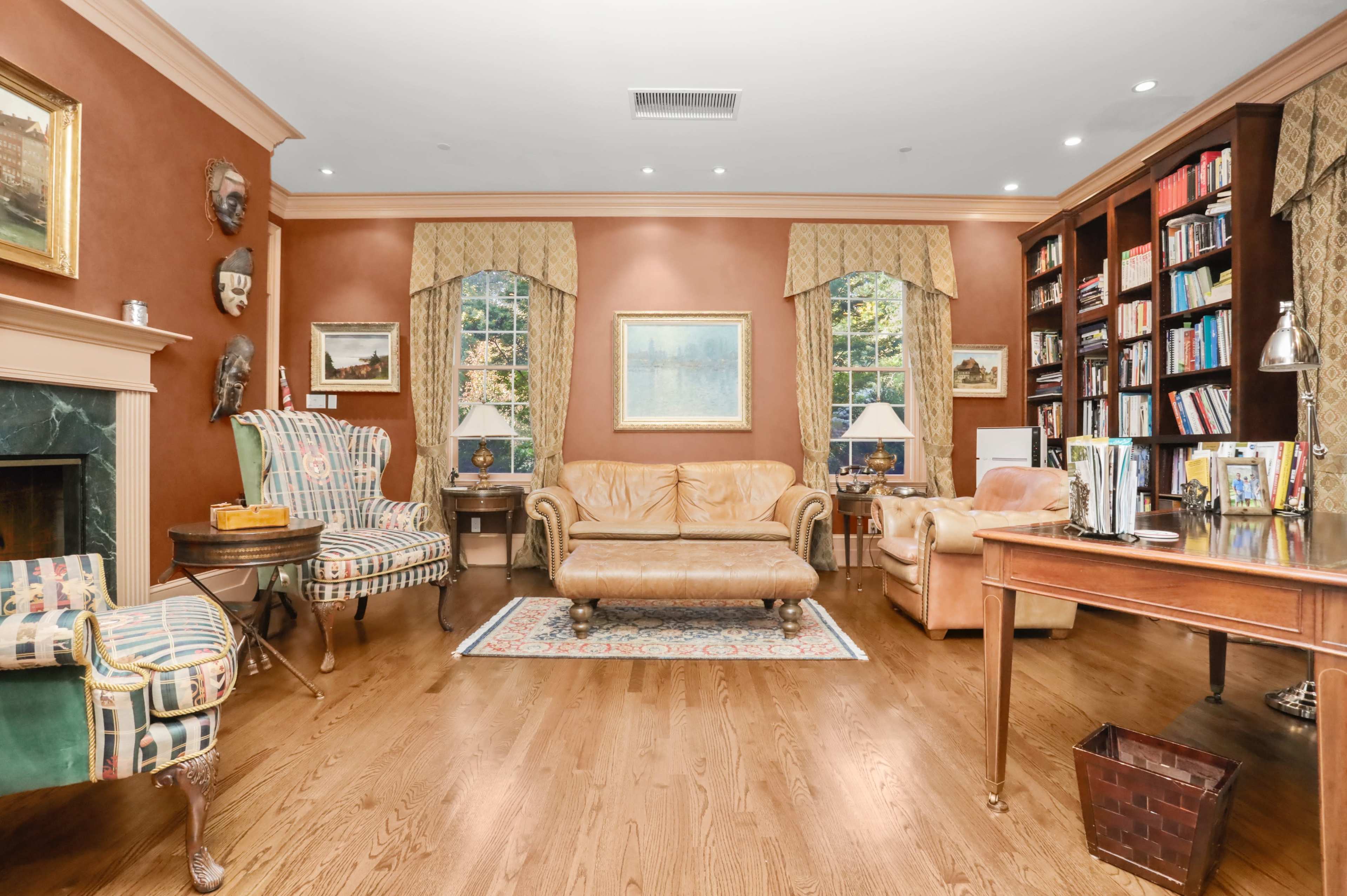A cozy living room featuring a leather sofa, two patterned armchairs, a wooden desk, and bookshelves filled with books, all set against a rich brown wall.