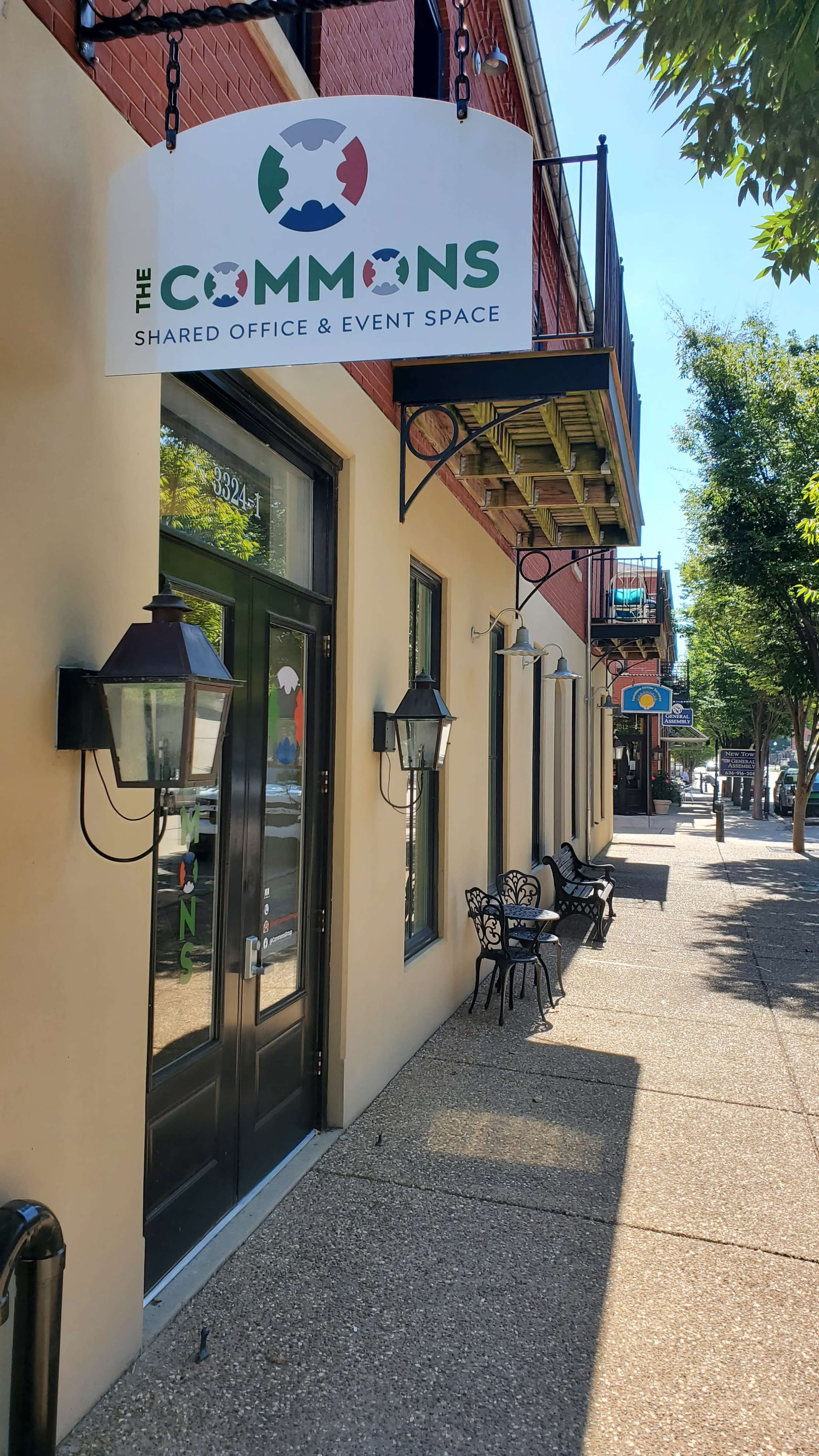 The image shows a street view of a building with a sign reading "The Commons," indicating a shared office and event space, and features outdoor seating along the sidewalk.