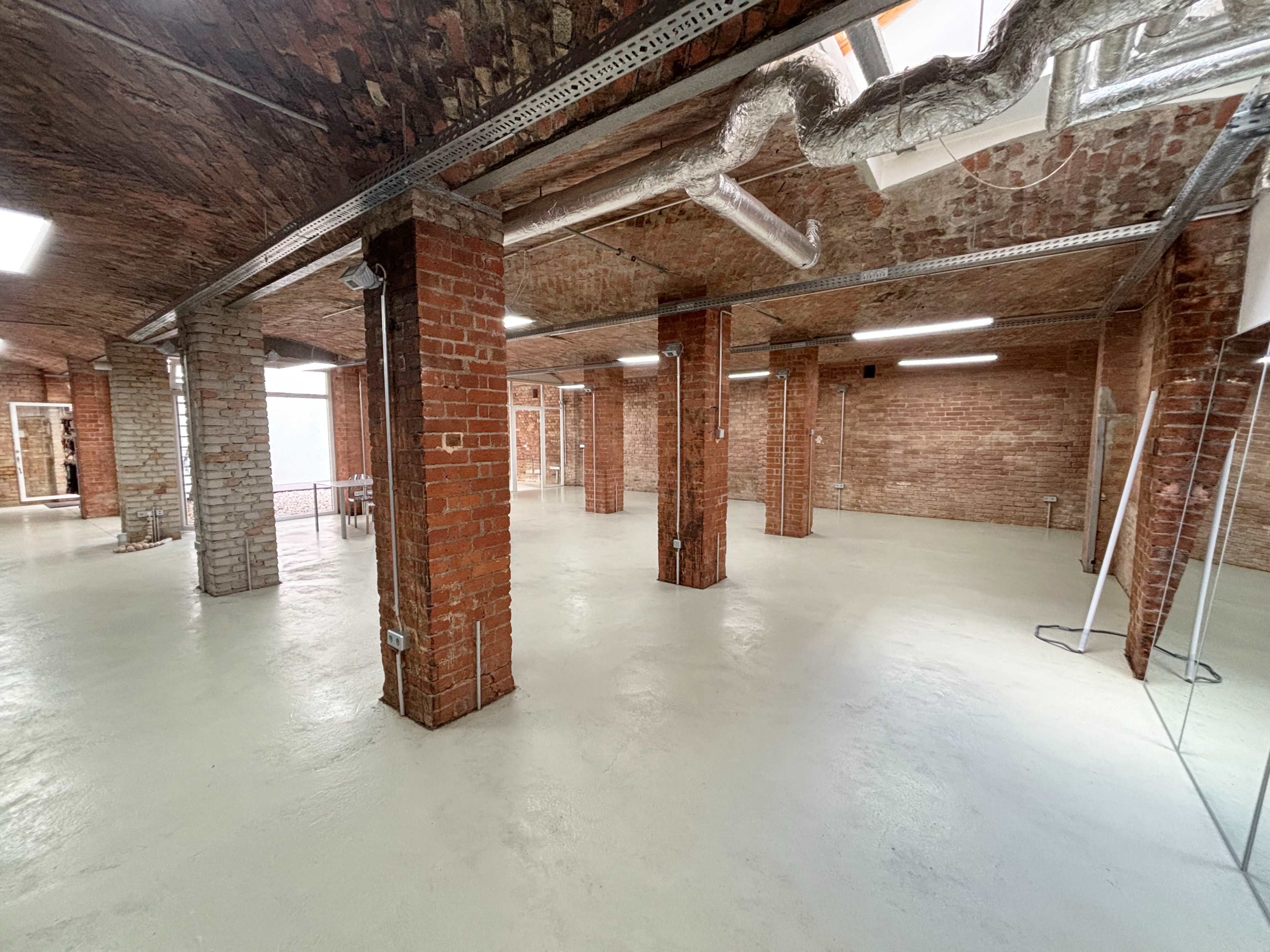 The image shows an empty industrial space with exposed brick walls, concrete floors, and several support columns.