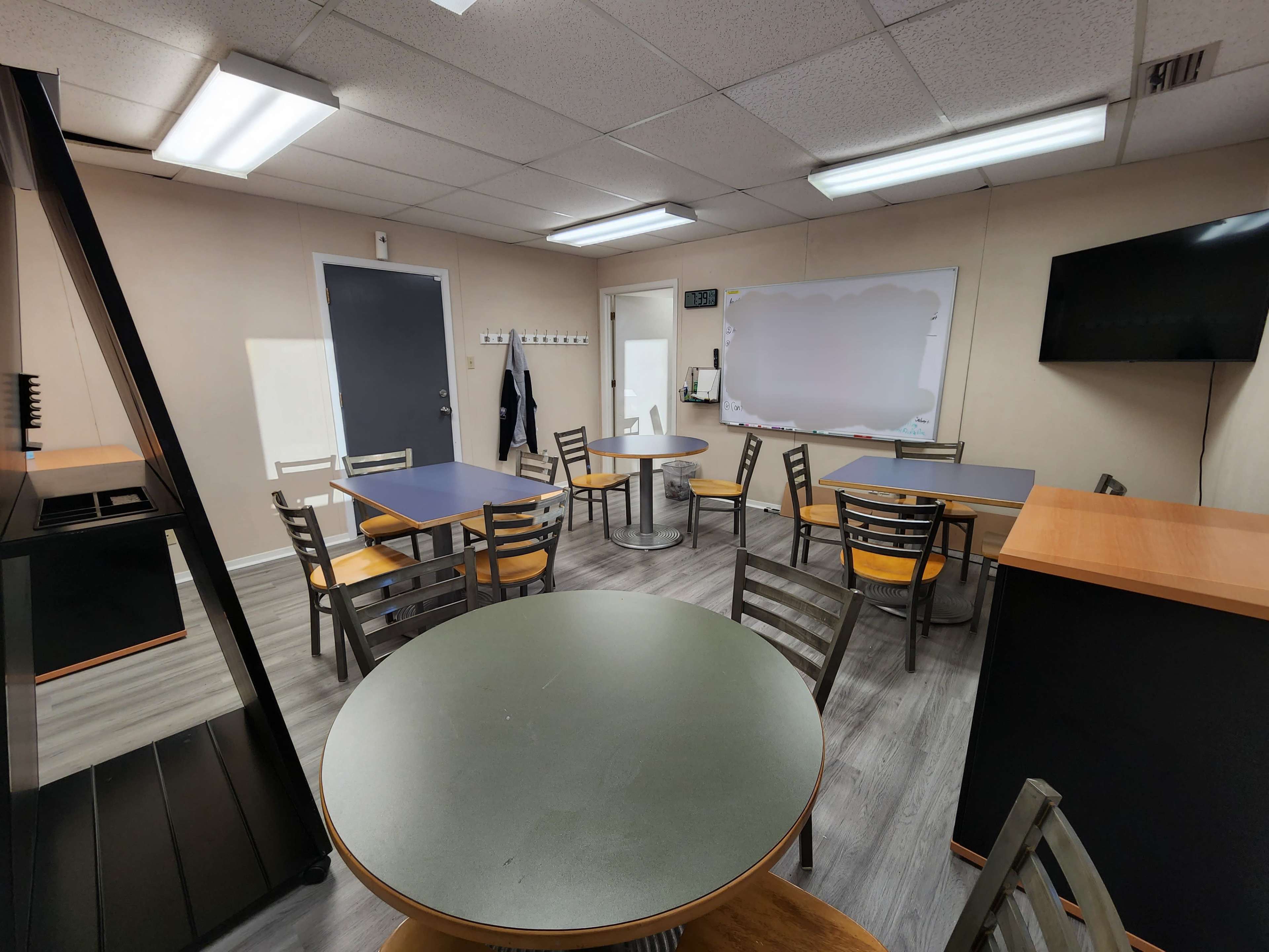 A sparsely furnished break room with tables and chairs arranged around the space, featuring a whiteboard, a television, and a door leading to another area.