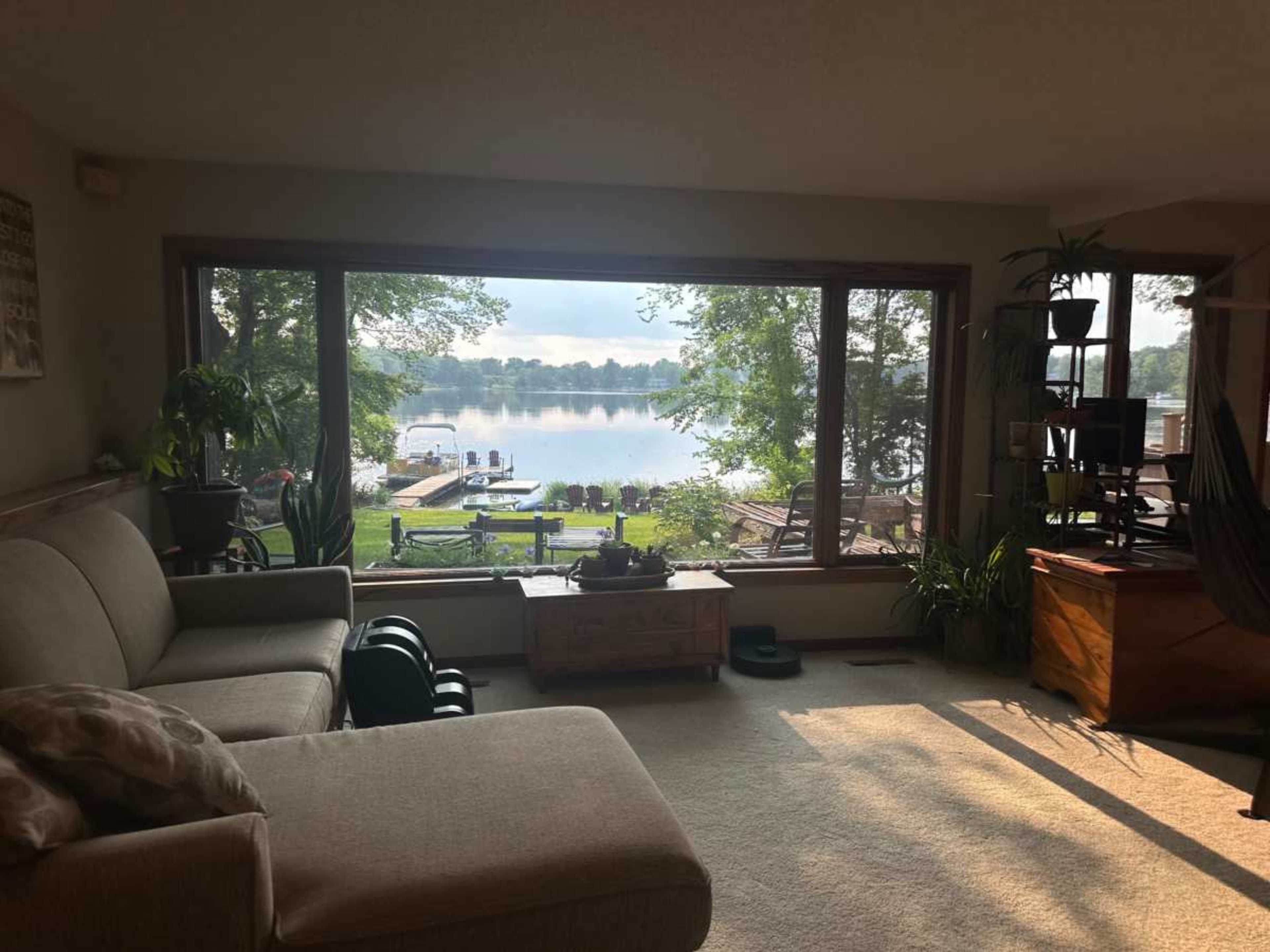 A cozy living room features a large window overlooking a serene lake surrounded by greenery and outdoor seating.