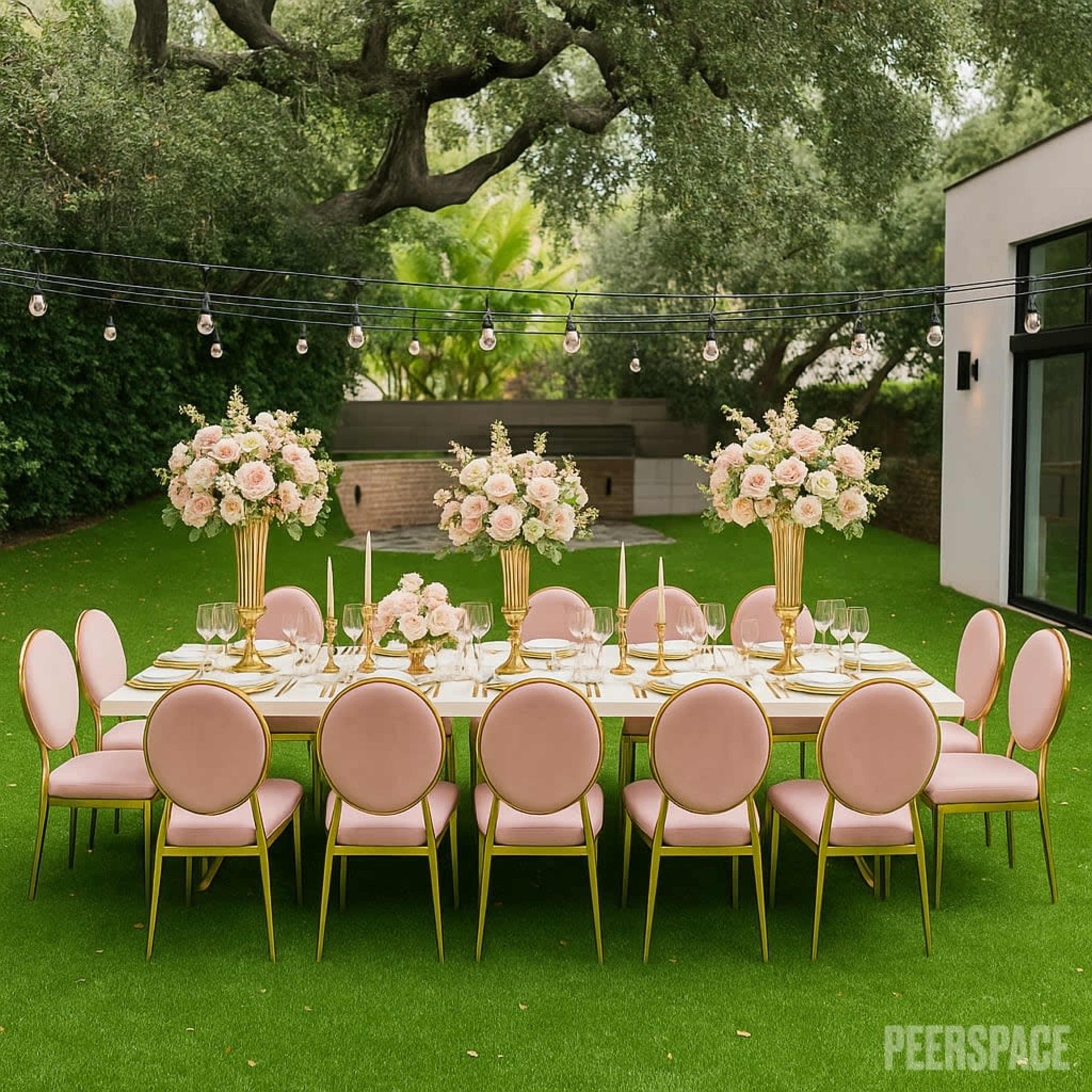 A long, elegantly set outdoor dining table with pink chairs and floral centerpieces is surrounded by greenery.