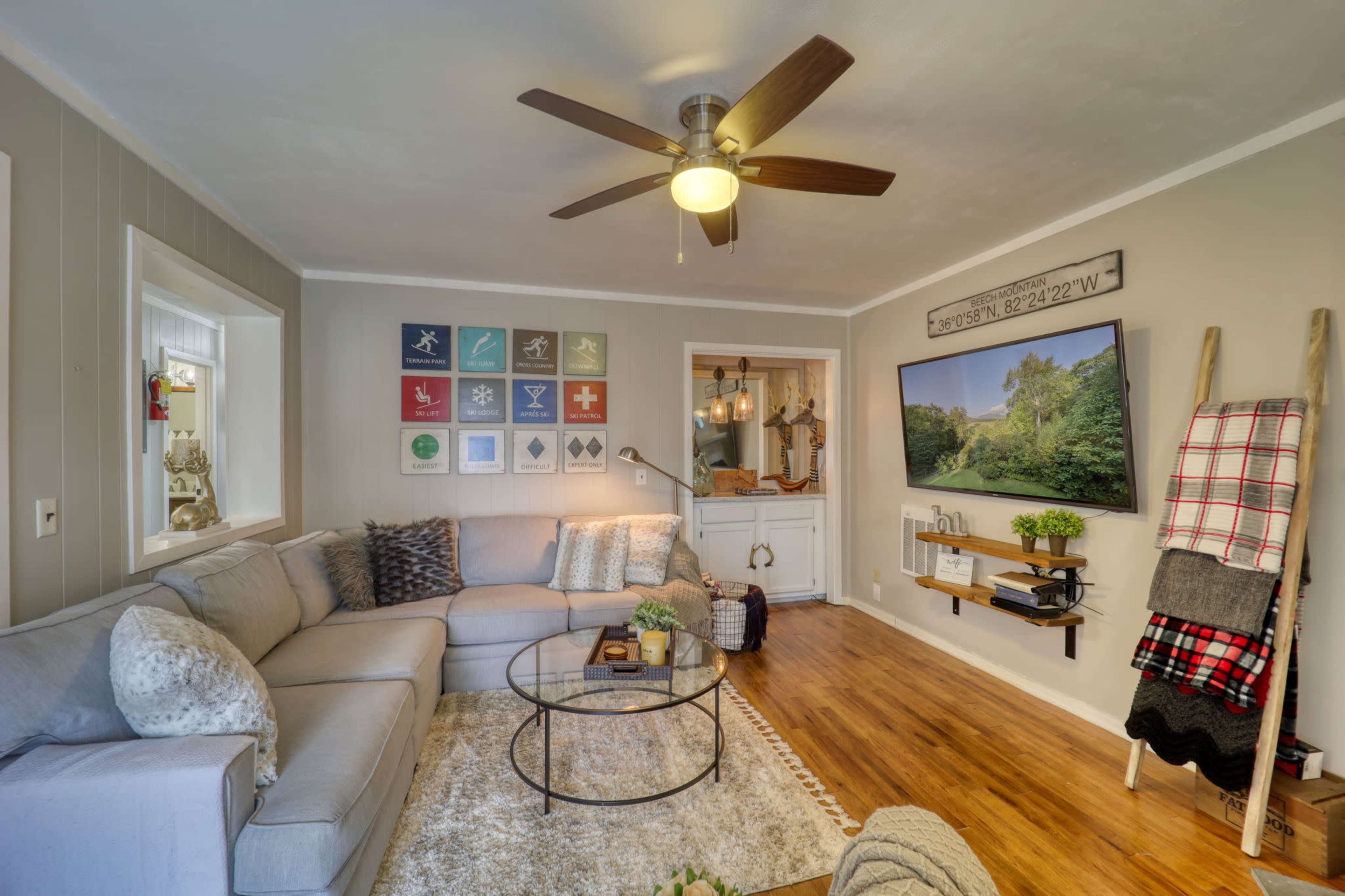 The living room features a light-colored sectional sofa, a round coffee table, wall art organized in a grid, and a flat-screen TV mounted above a wooden shelf.