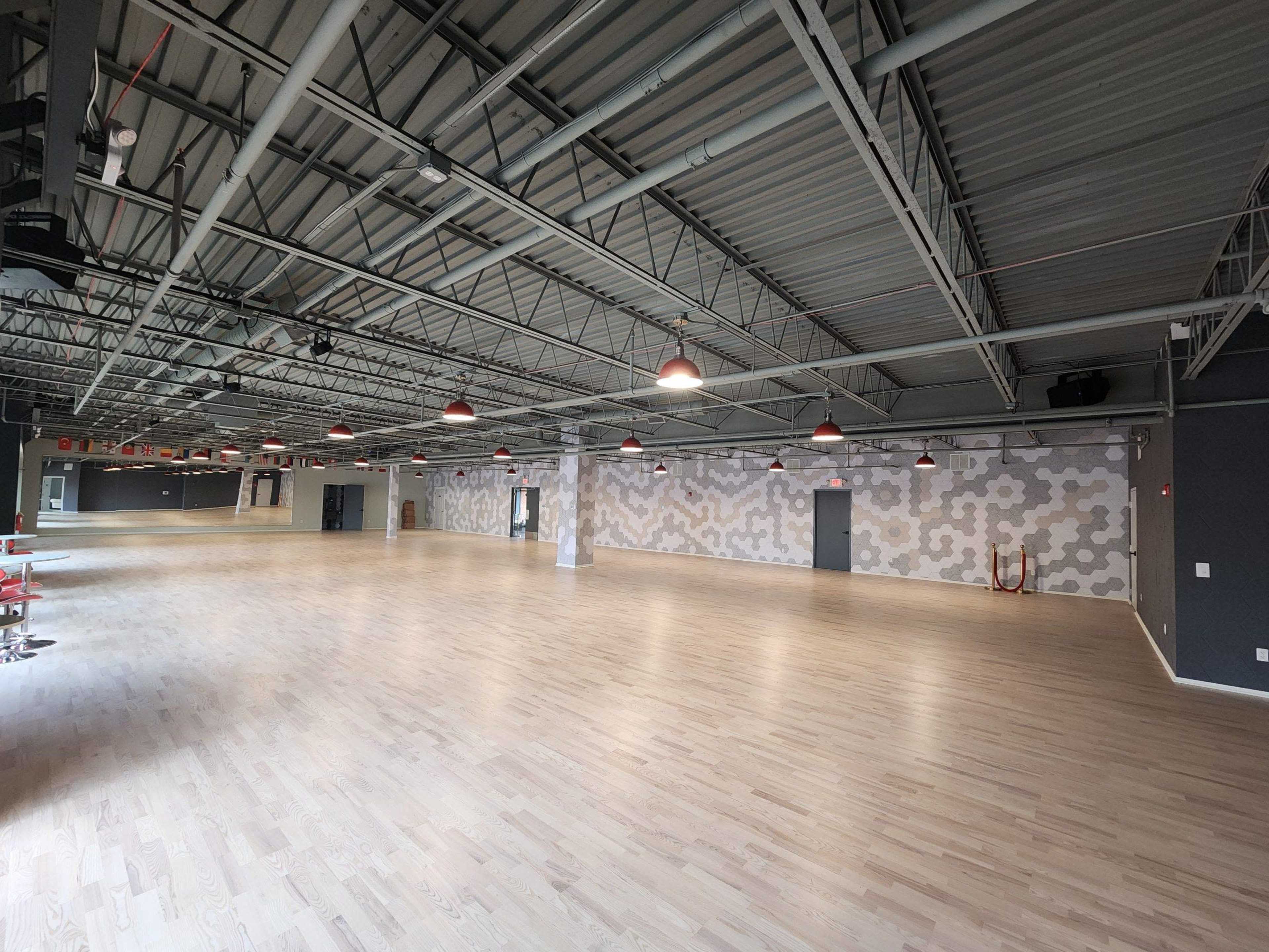 The image shows a spacious, empty interior with wooden flooring, industrial-style lighting, and patterned walls.