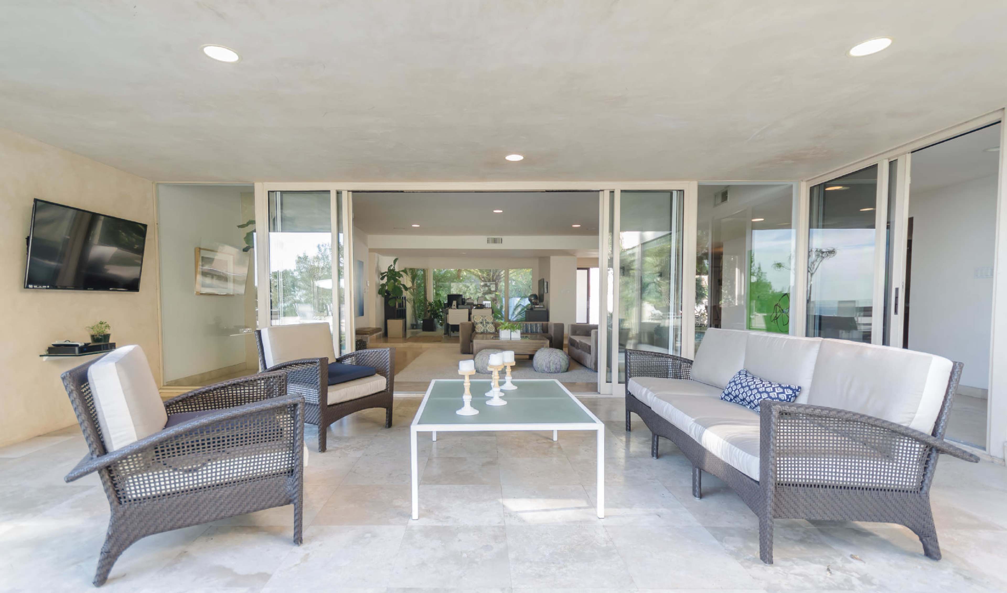 A spacious outdoor seating area features wicker furniture arranged around a coffee table, with a view into a bright interior space through large glass doors.
