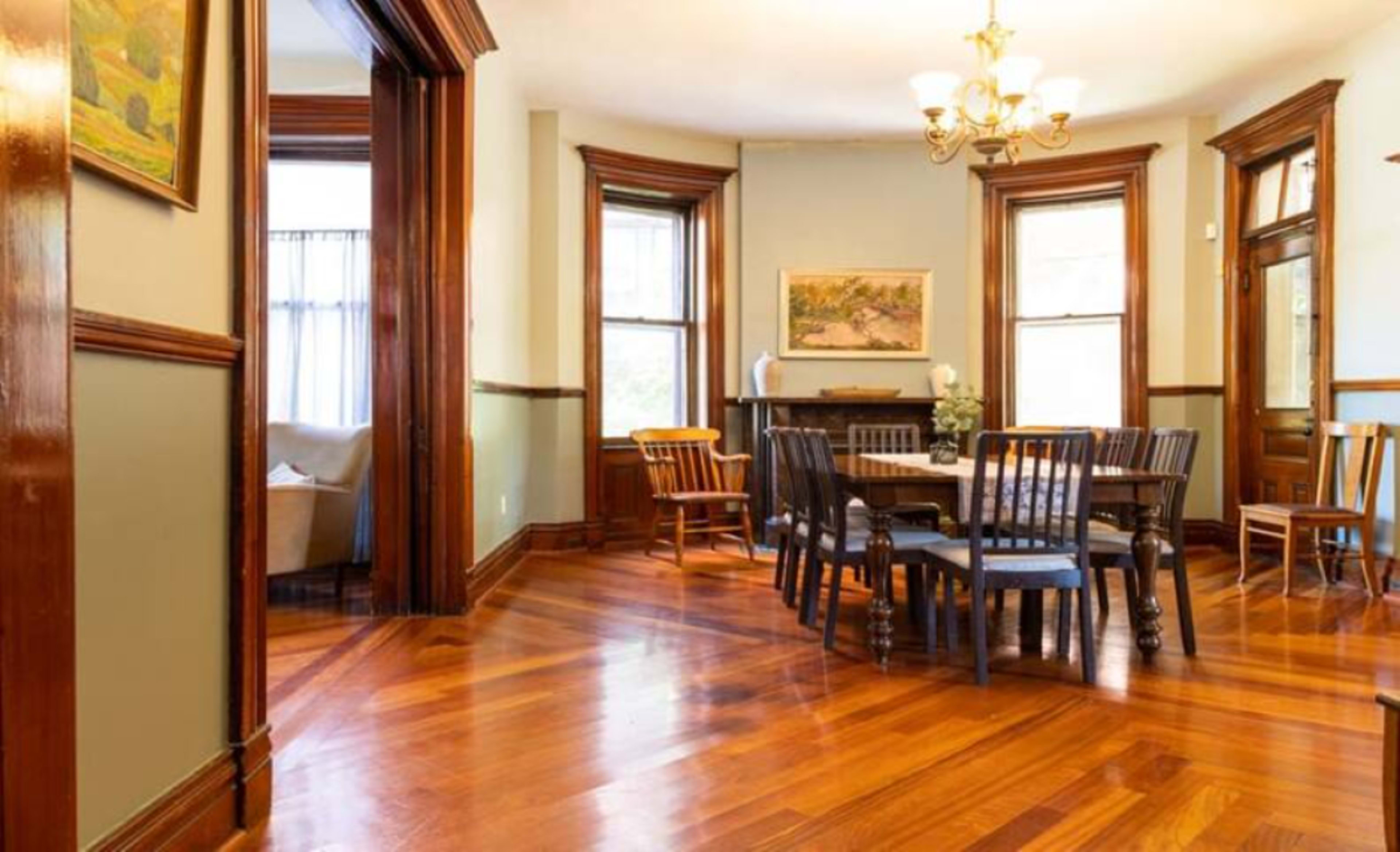 The image shows a dining area with wooden flooring, a table surrounded by chairs, and windows allowing natural light to enter the room.