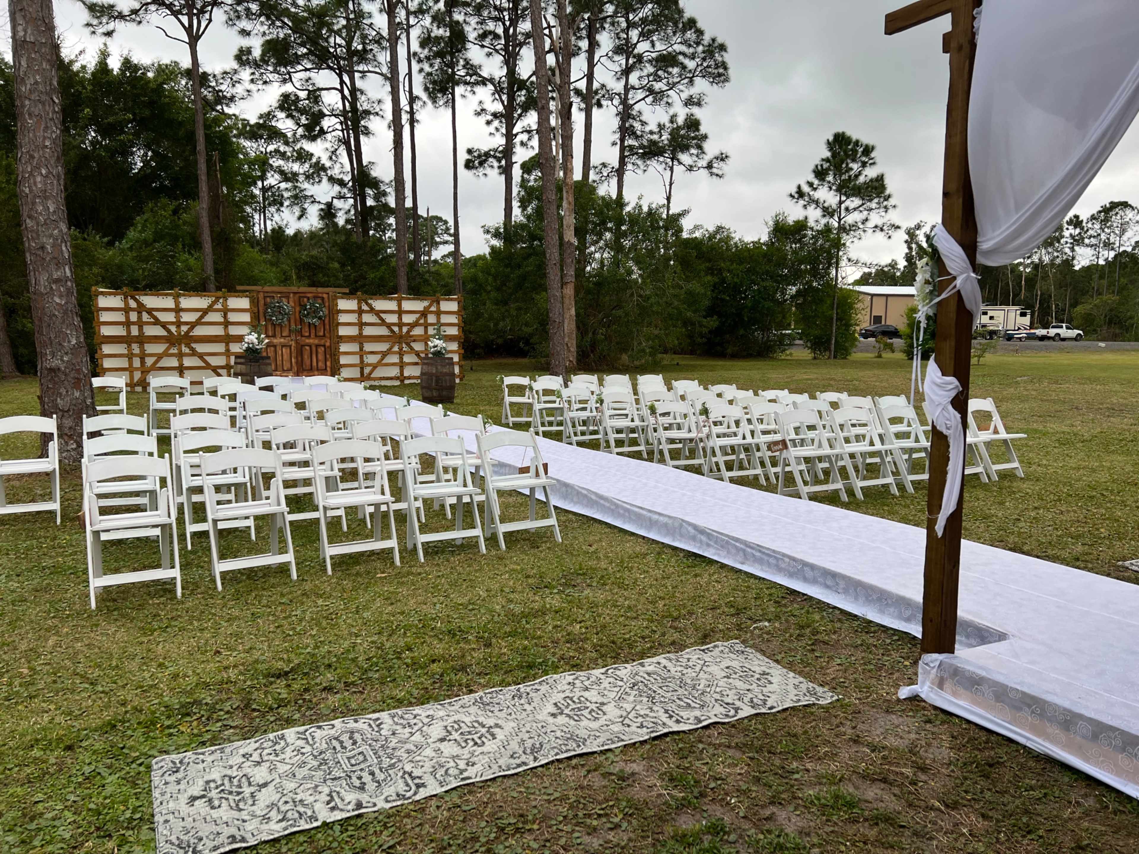 The image shows an outdoor wedding setup with rows of white chairs facing a decorated wooden arch and a white carpet leading to it, surrounded by trees and a grassy area.