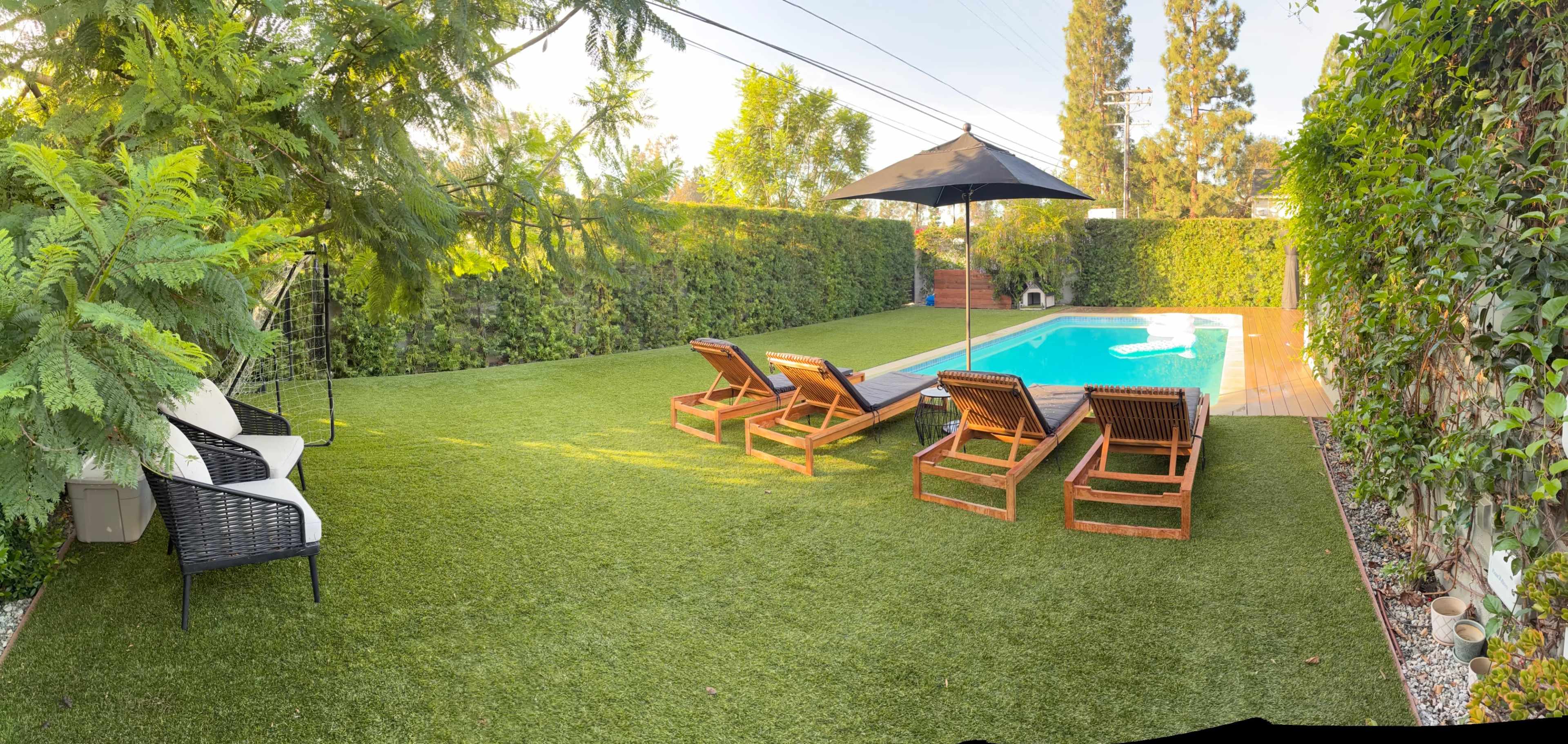 The image shows a landscaped backyard with a swimming pool surrounded by lounge chairs and a shaded area, featuring lush greenery and artificial turf.