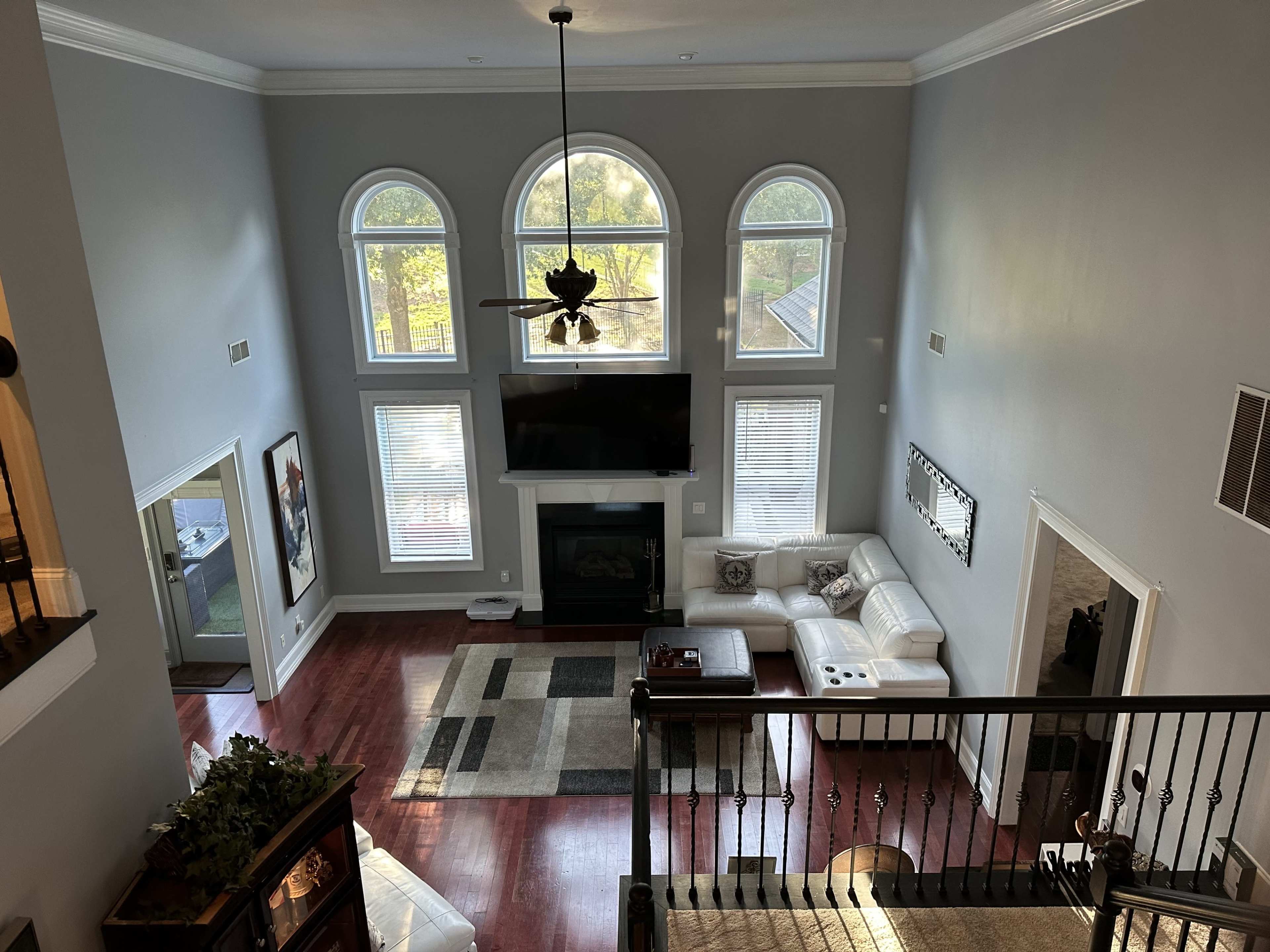 The image shows a spacious living room with large windows, a flat-screen TV mounted above a fireplace, and modern furniture arranged around a central area rug.