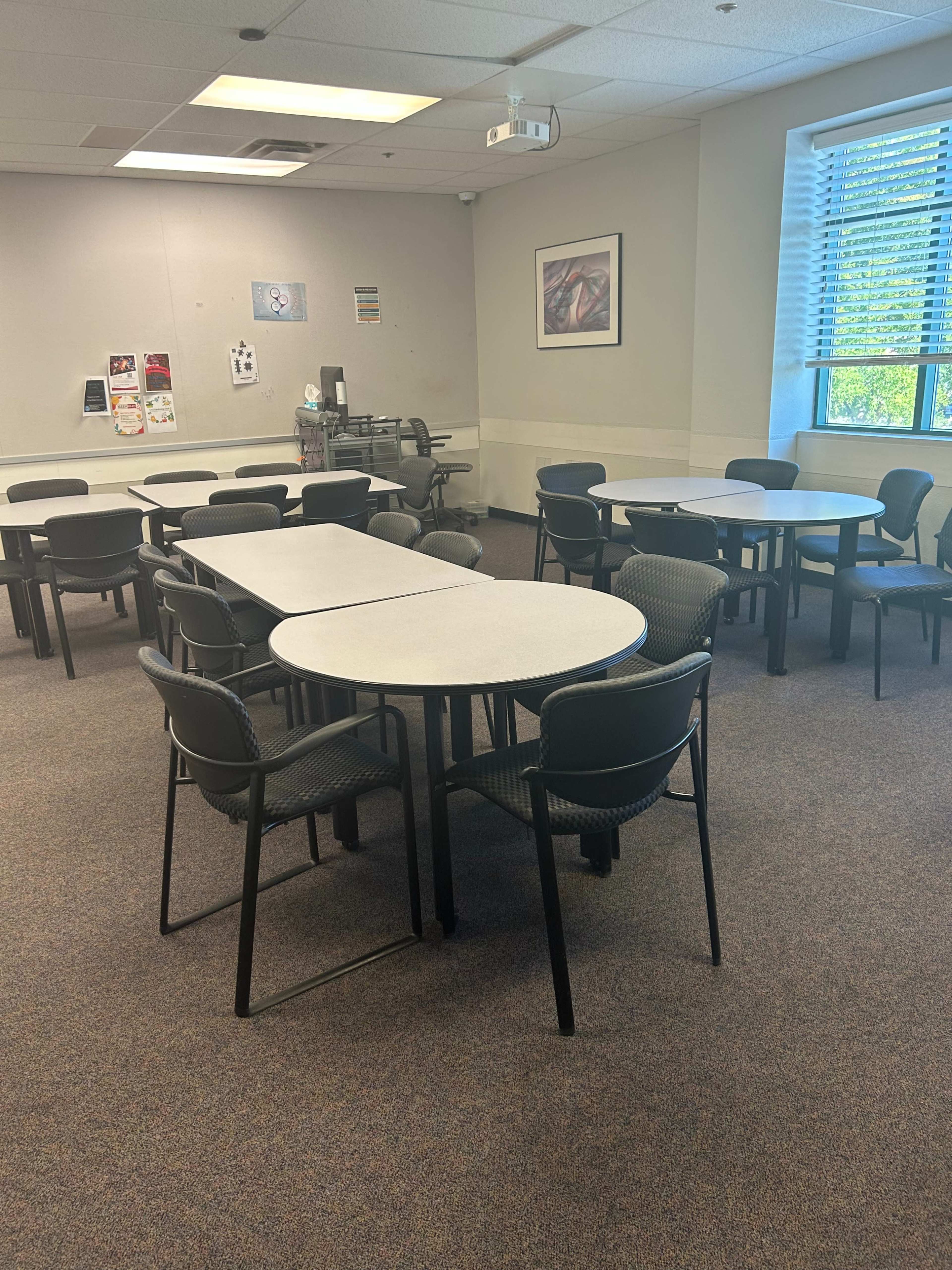 A classroom or meeting room with multiple round tables and chairs arranged in a spacious layout.