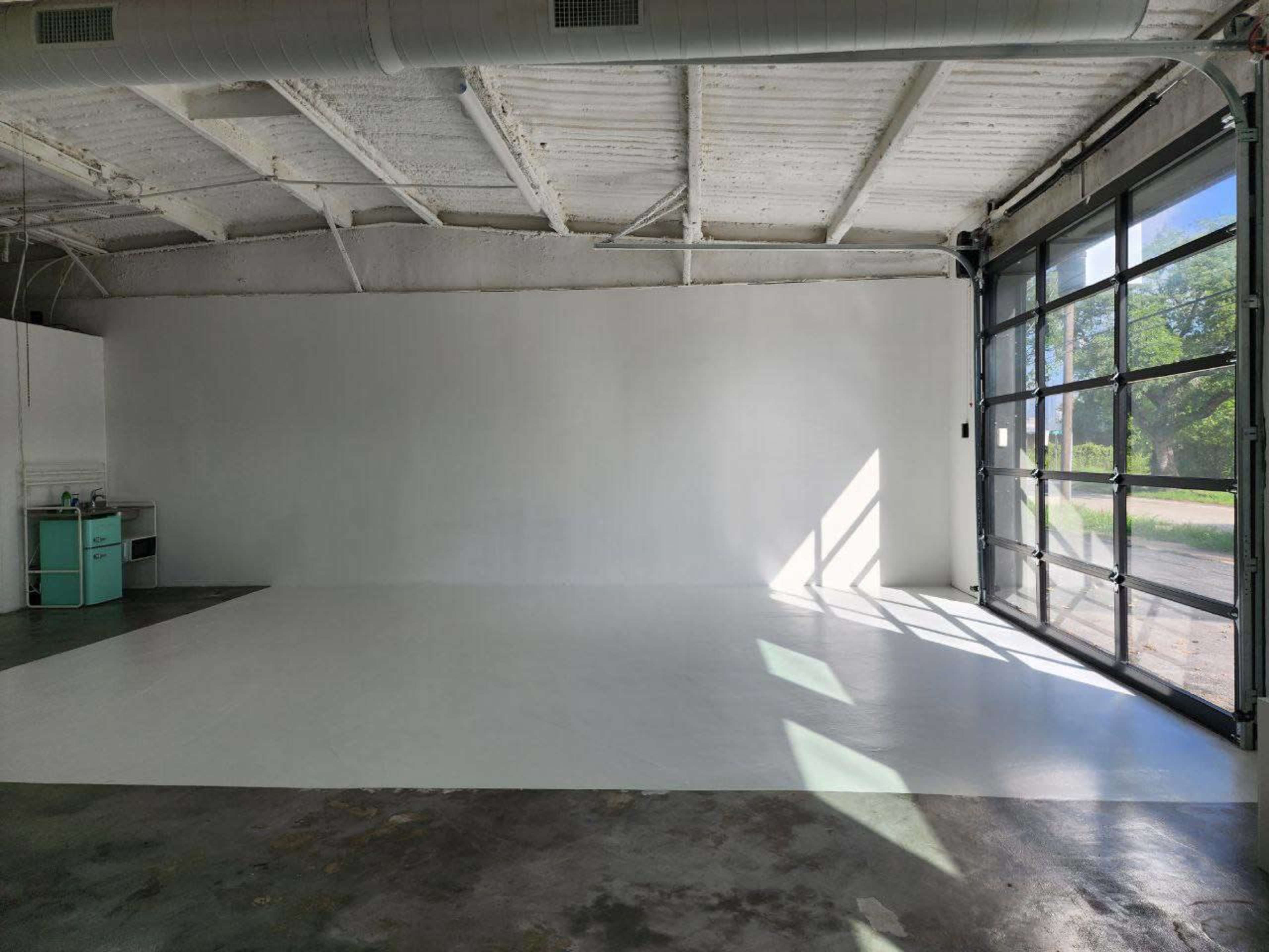 An empty room with a white floor and walls, featuring a large garage door that opens to an outdoor view.