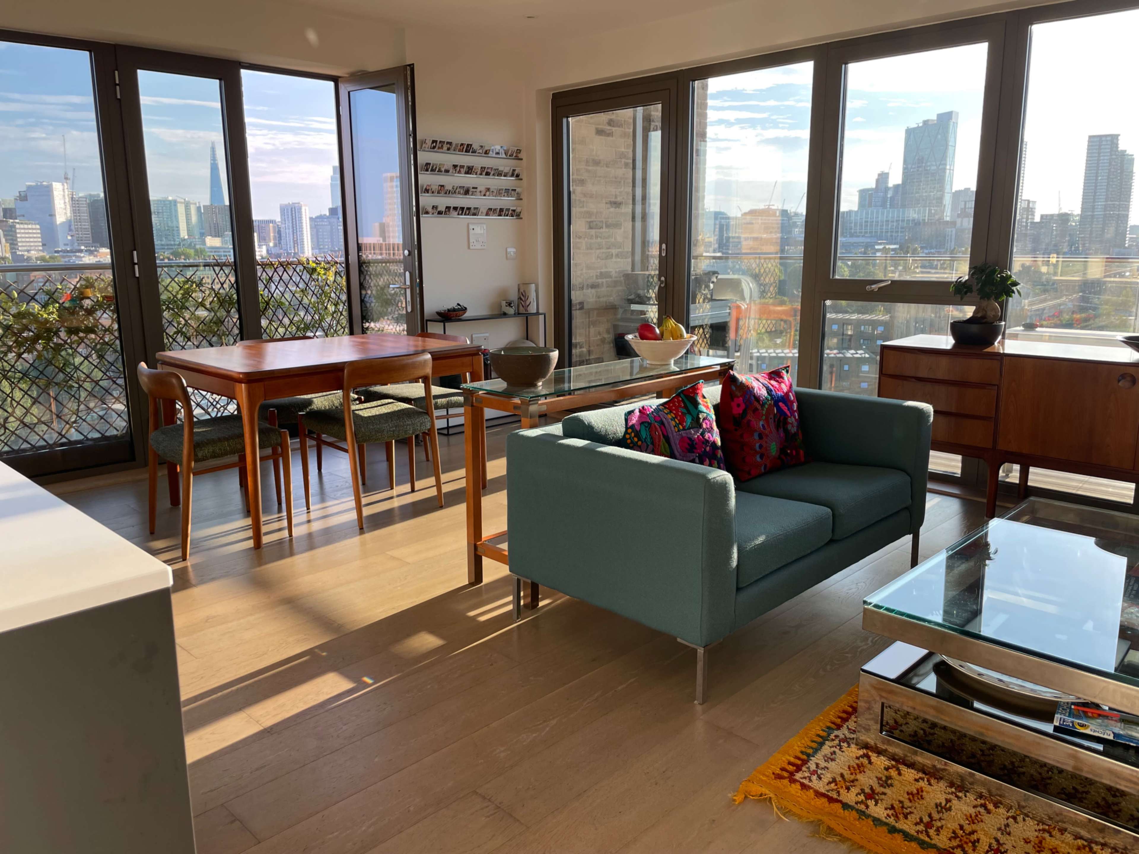 A bright, modern apartment features a teal sofa, a glass coffee table, a wooden dining table, and large windows overlooking a cityscape.