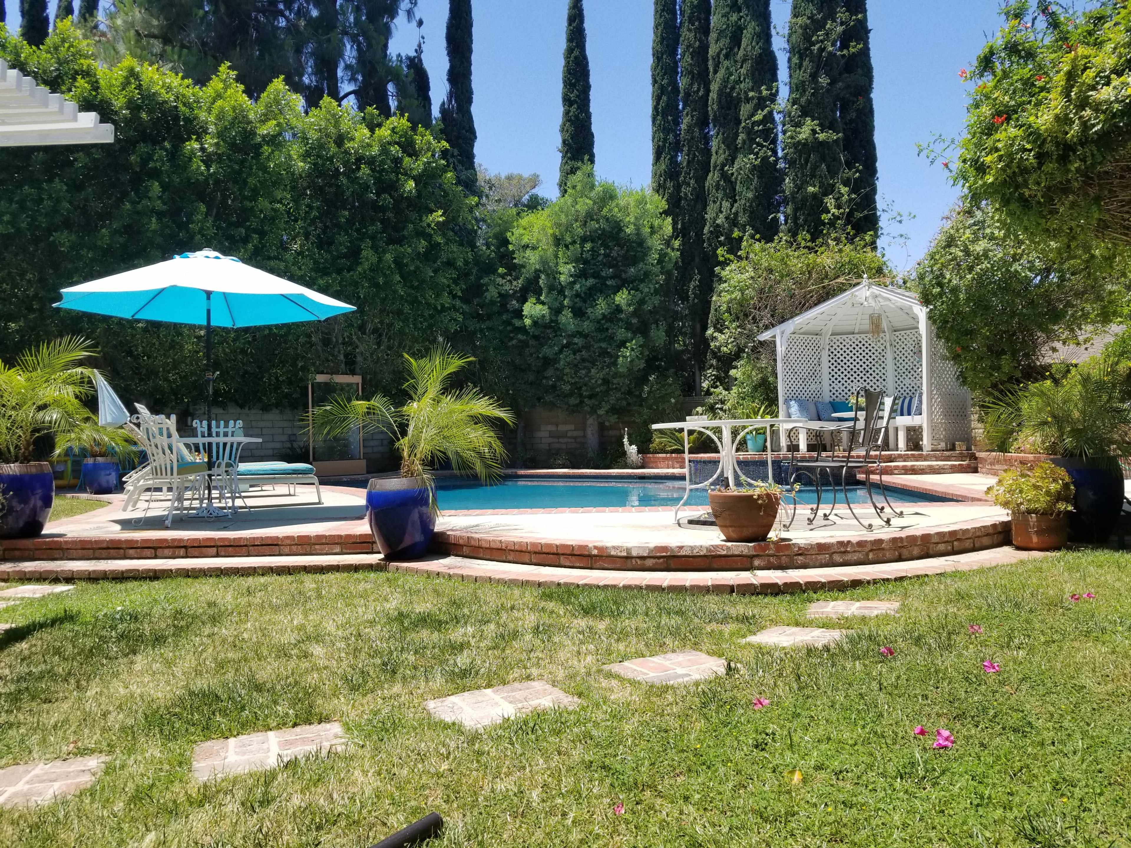 A backyard features a swimming pool surrounded by green grass, a patio with outdoor furniture, and tall trees in the background.