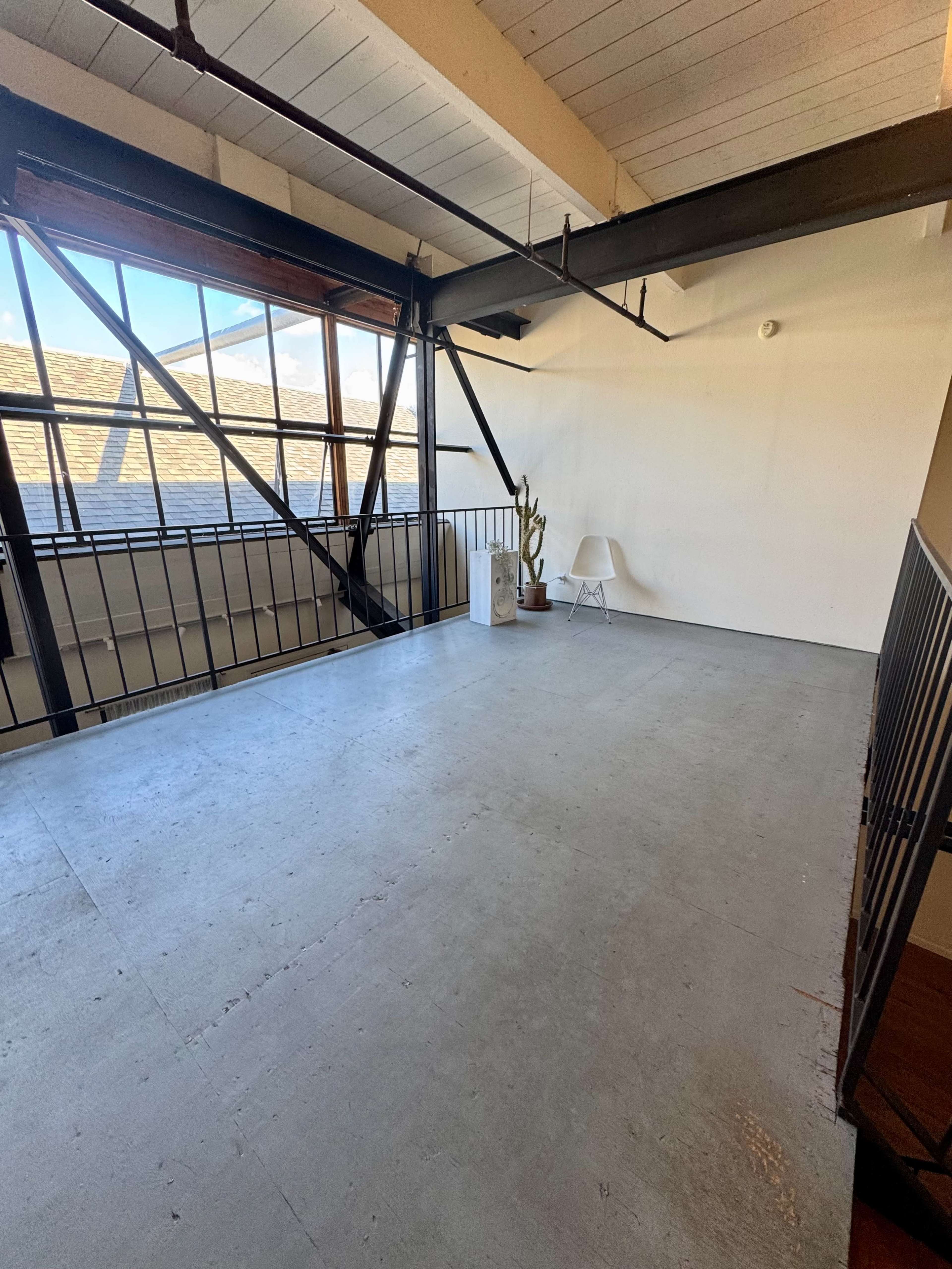 The image shows a spacious, empty loft-style room with large windows and industrial beams.