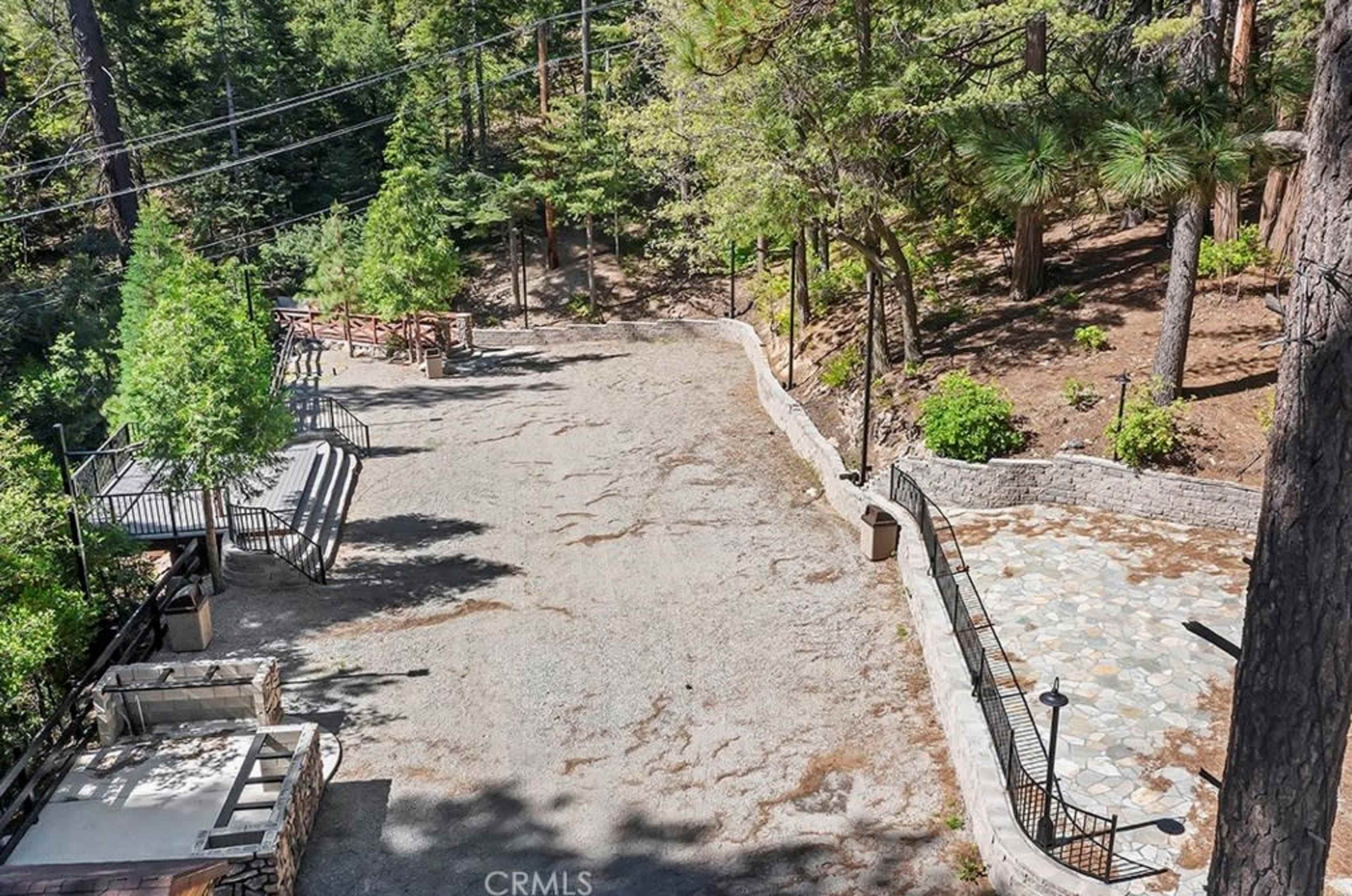 The image shows a spacious outdoor area surrounded by trees, featuring a gravel surface and a stone pathway, with some seating and fencing nearby.