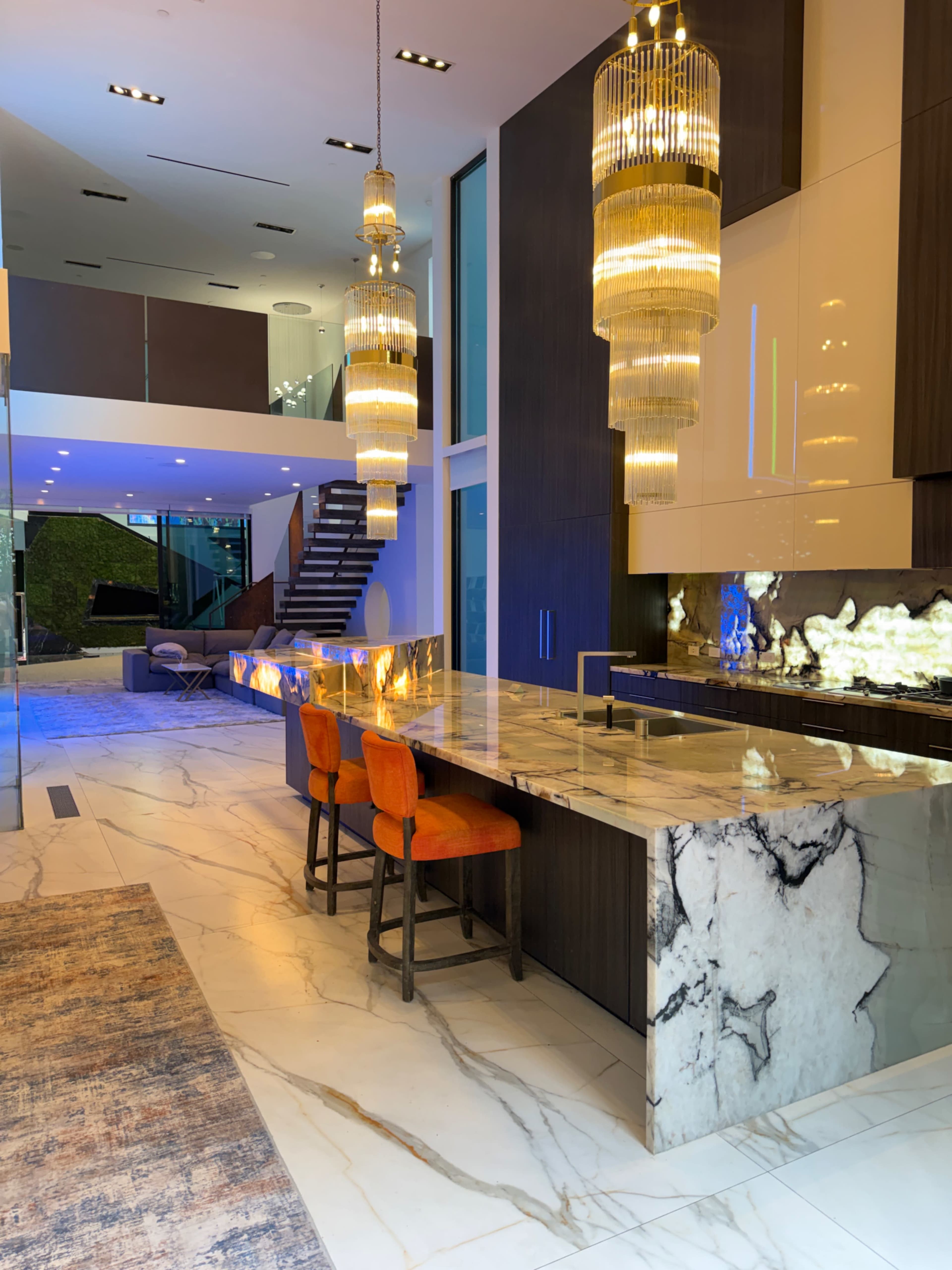 The image shows a modern kitchen with a marble island, four orange bar stools, and two large, decorative light fixtures hanging above.