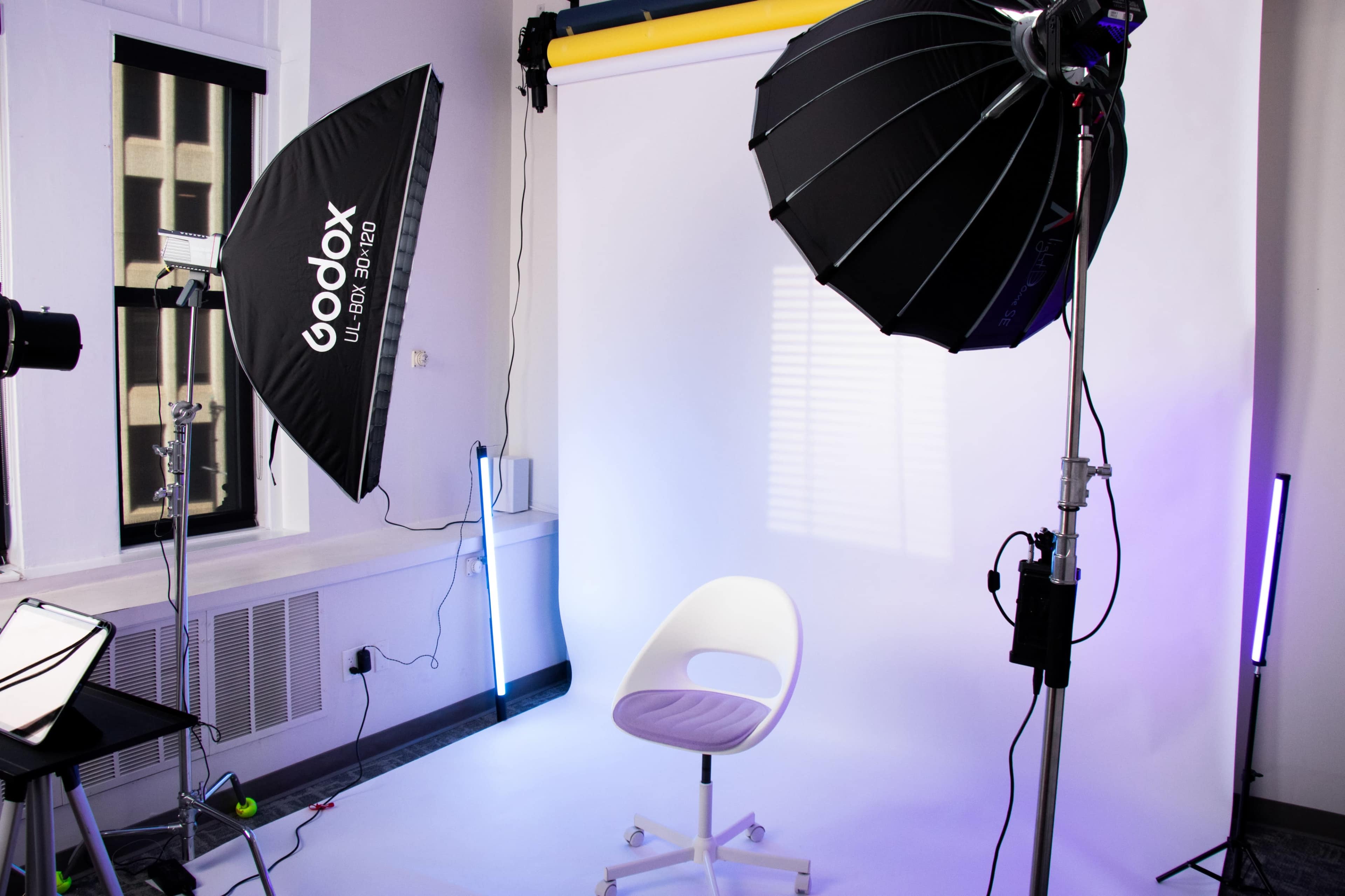 The image shows a photography studio setup with a white backdrop, a chair in the center, and multiple lighting equipment including softboxes and a light stand.