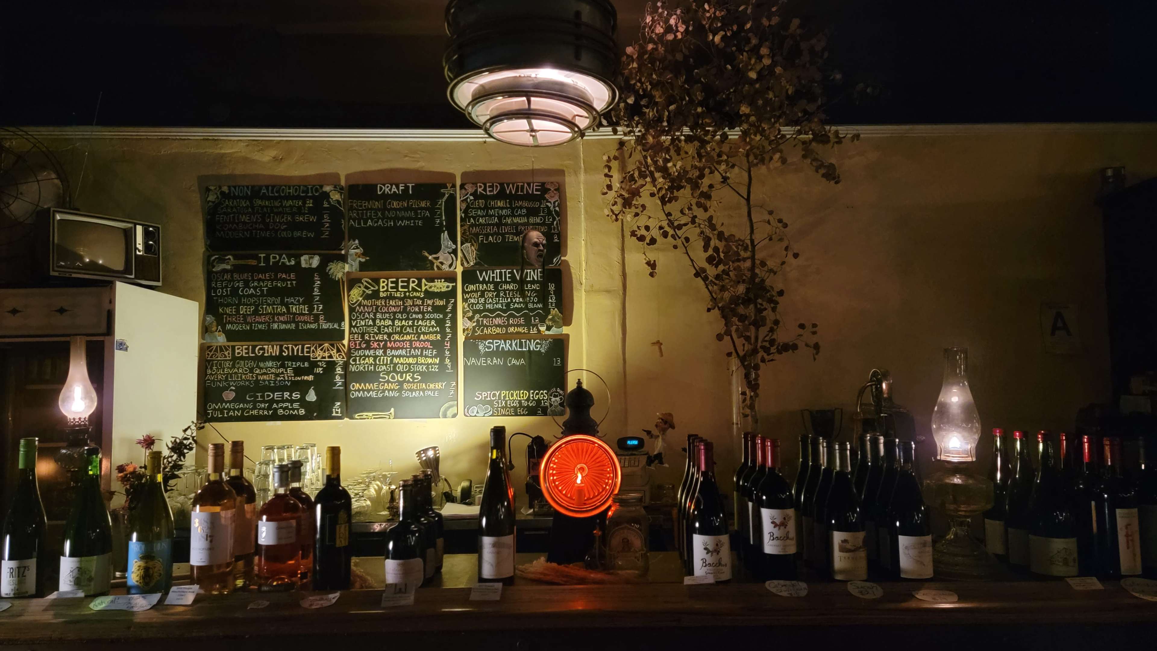 A dimly lit bar displays a collection of wine bottles and a chalkboard menu featuring various drinks.