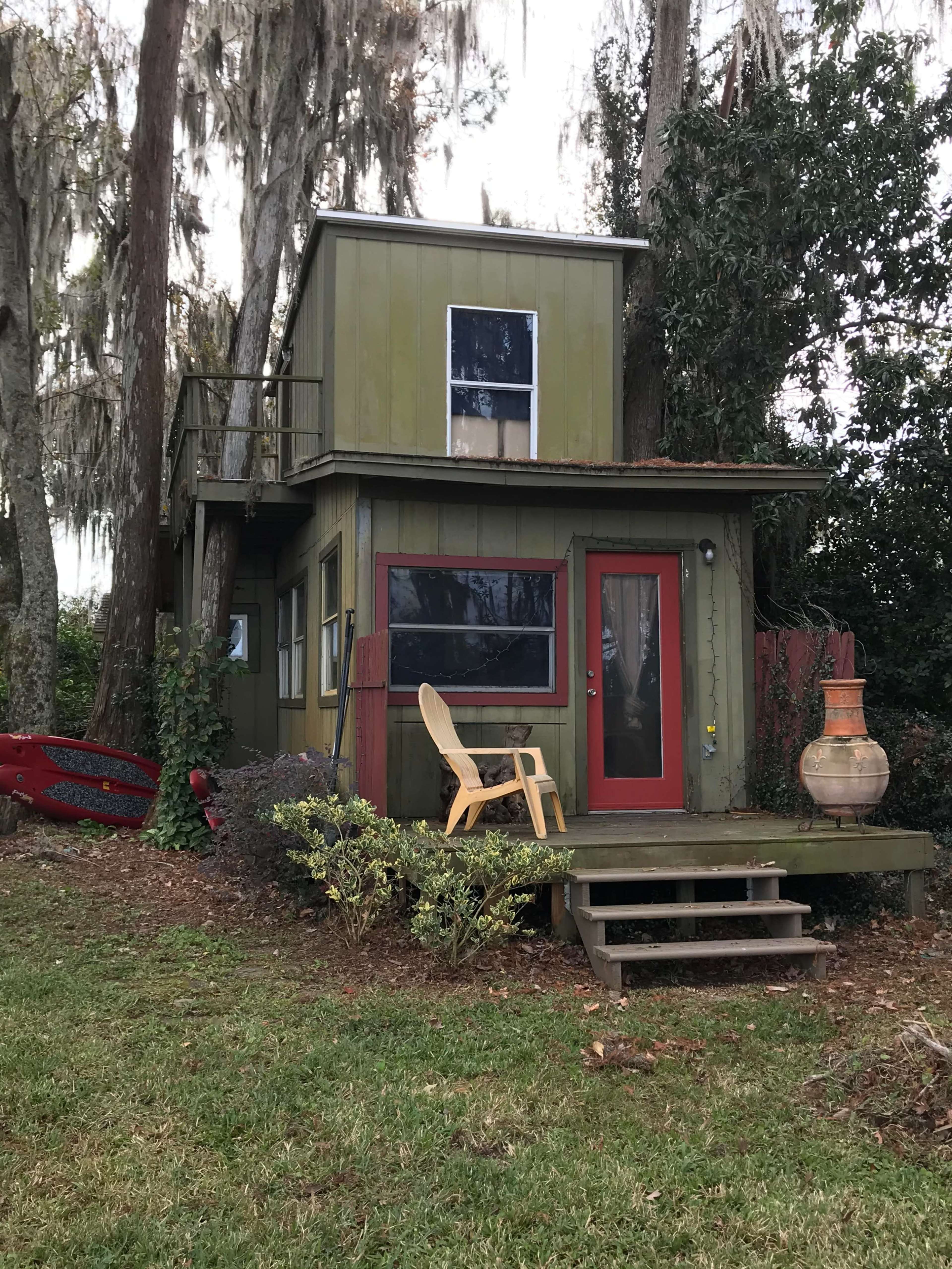 A small two-story green house with a red door and a wooden deck is surrounded by trees and grass.
