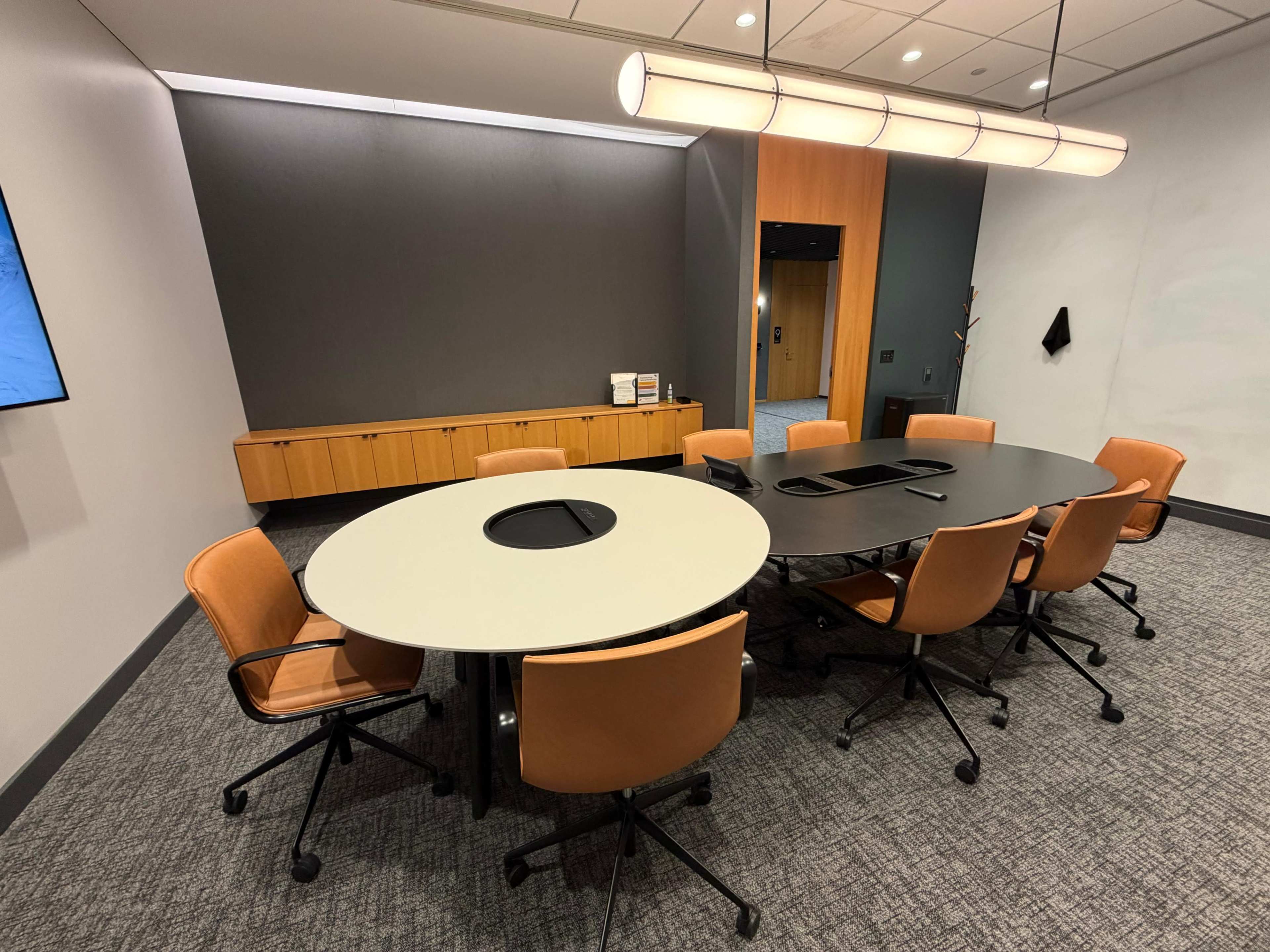 A modern conference room features a large round table surrounded by several orange office chairs, with a wall-mounted display and sleek cabinetry in the background.