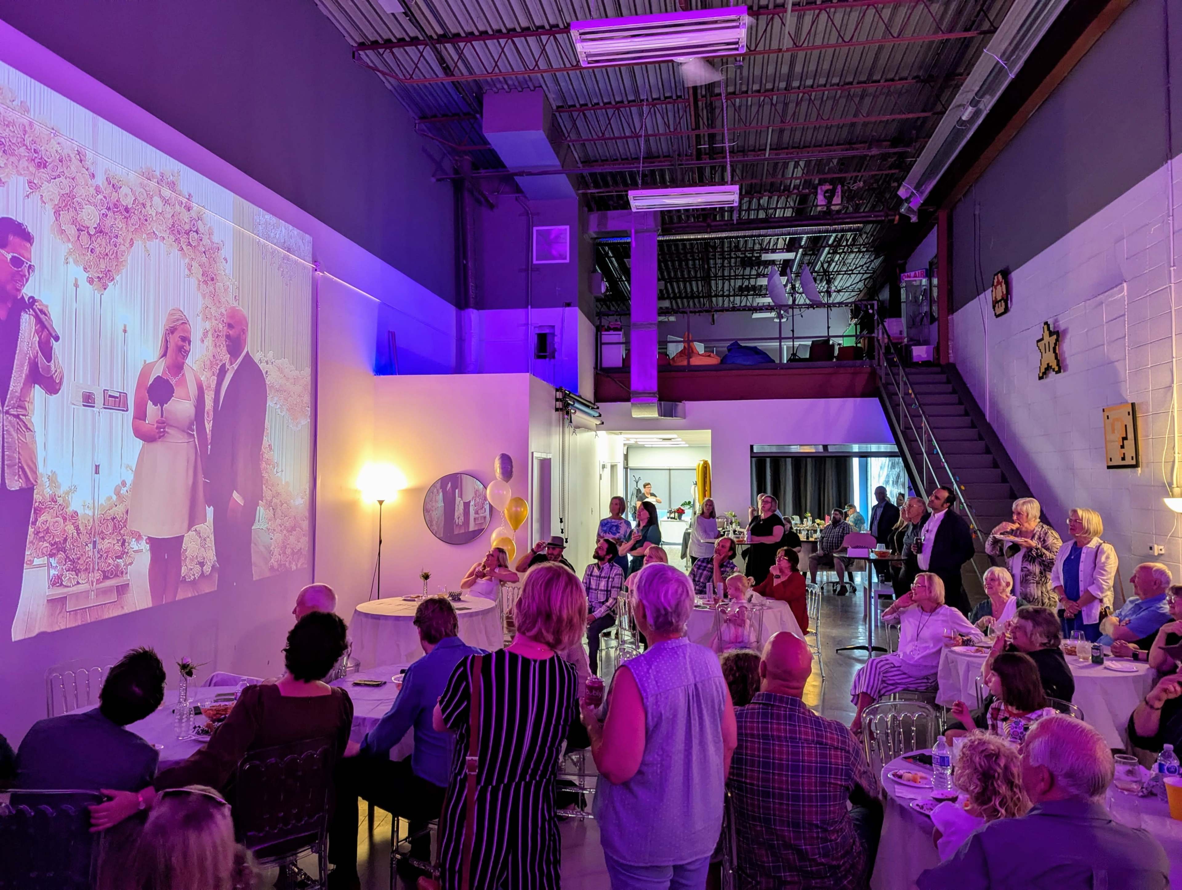 A group of people are seated and standing in a venue, watching a projected image of a couple on a screen during a celebration event.