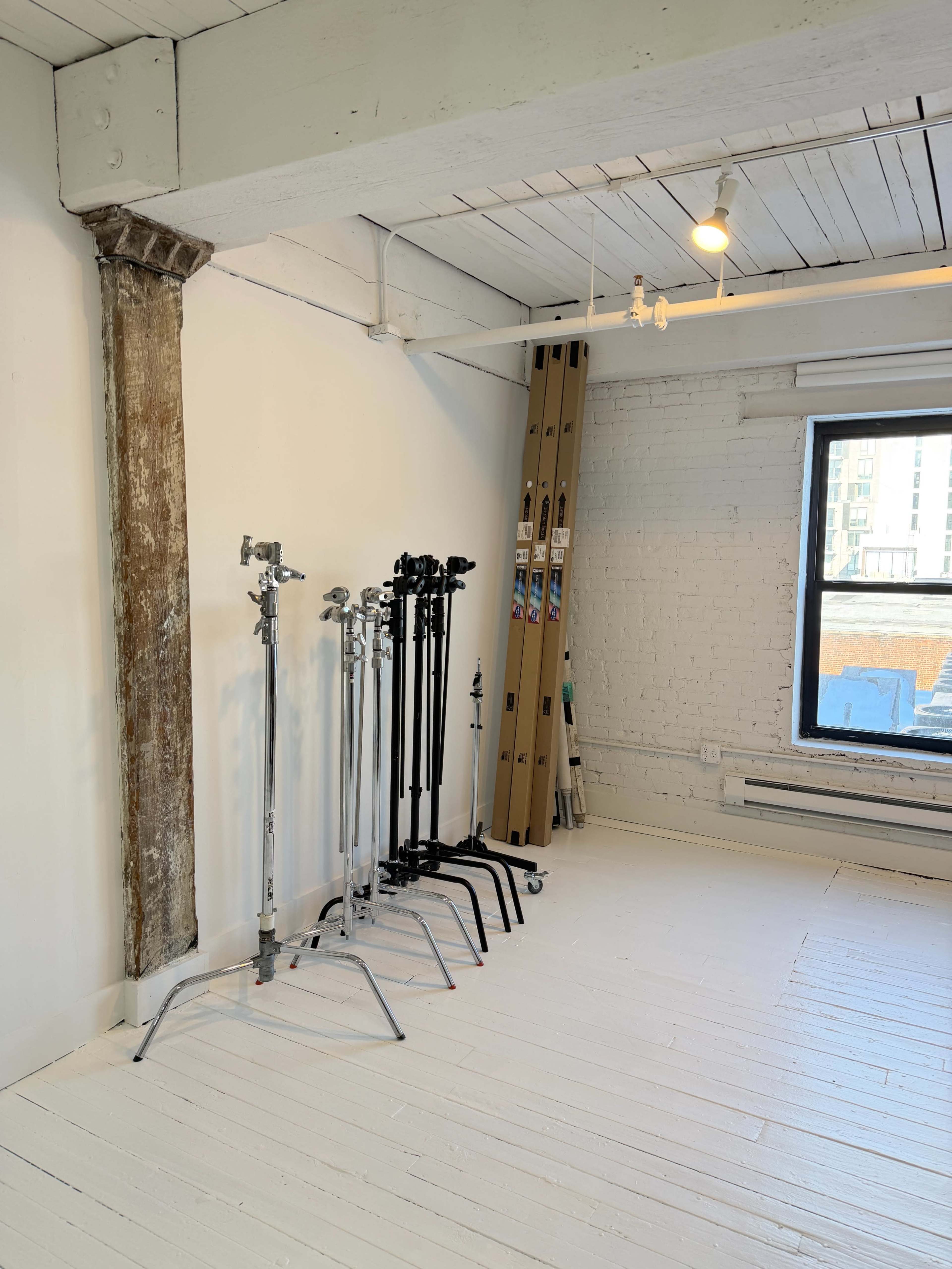 The image shows a bright, spacious room with a brick wall, a wooden ceiling, and several lighting stands arranged neatly against one wall.