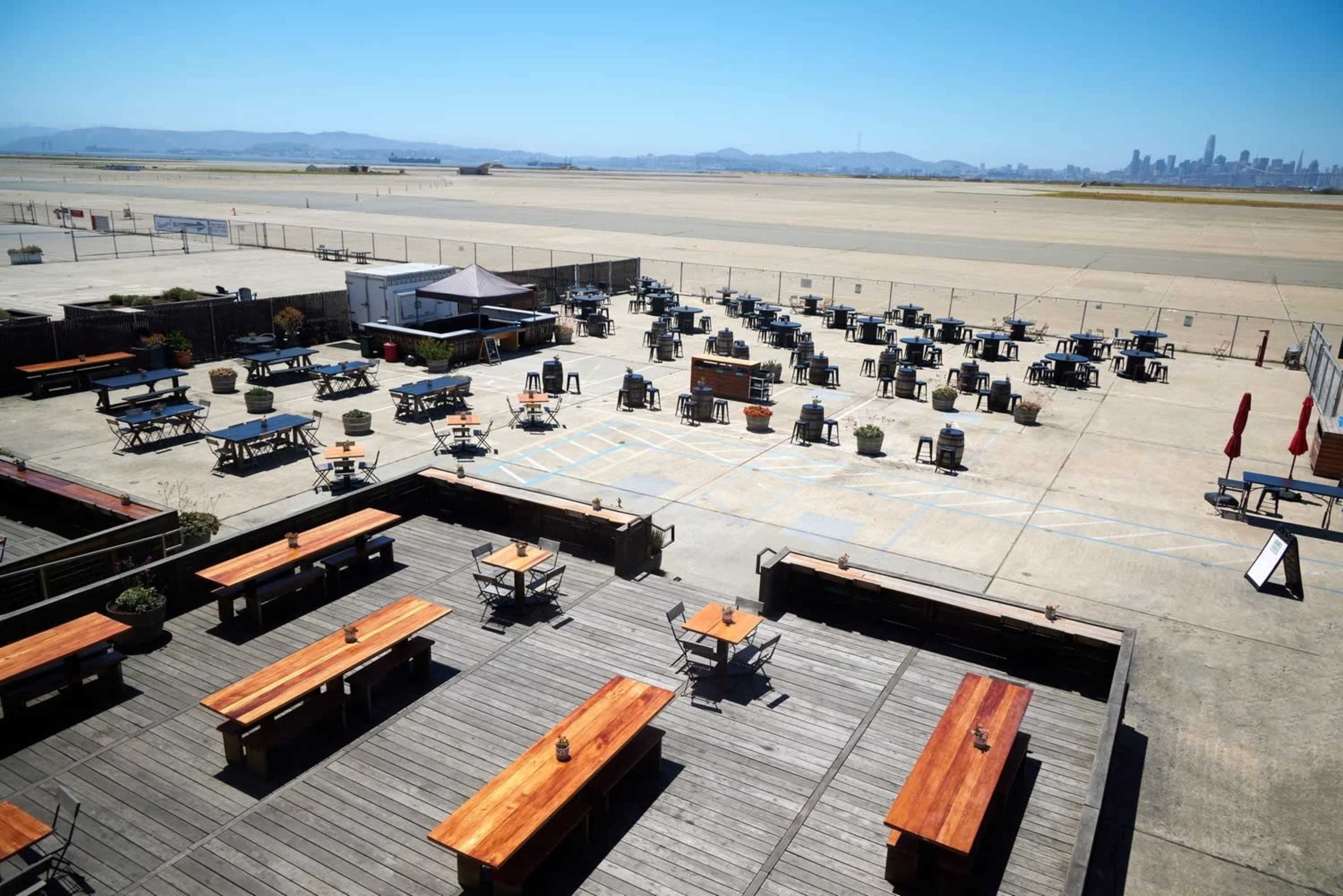 An outdoor seating area with numerous wooden tables and chairs is set up on an expansive concrete space next to an empty runway.