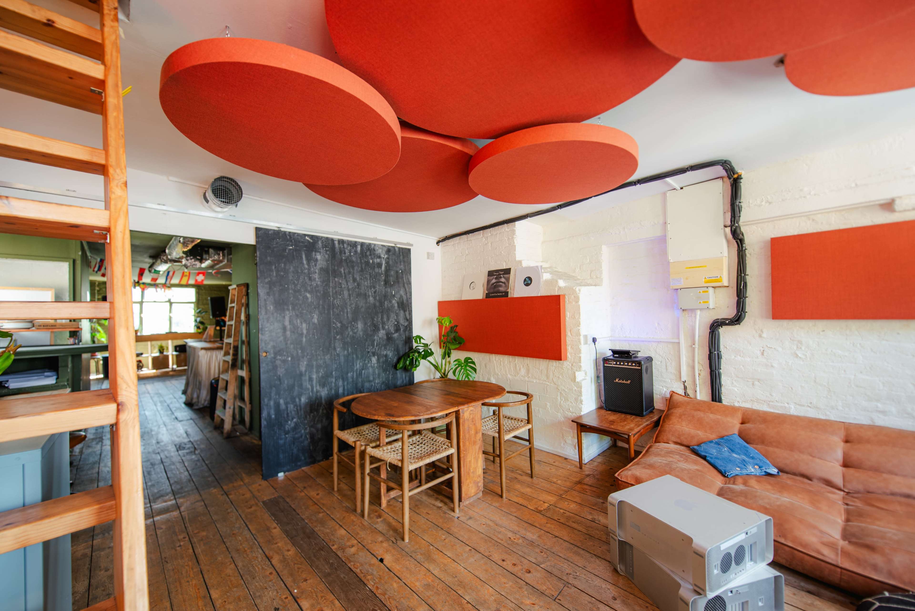 A cozy, modern living space features a wooden floor, a brown sofa, a round wooden dining table surrounded by woven chairs, and acoustic panels on the ceiling and walls.